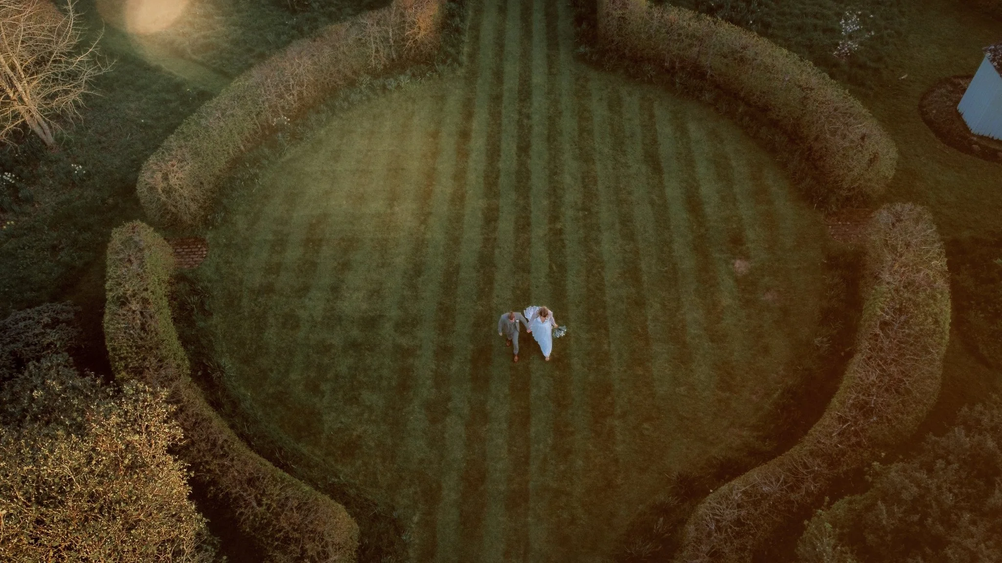 An aerial view of a bride and groom walking hand-in-hand across a grassy, decorated backyard with hedges and trees, illuminated by warm lighting.