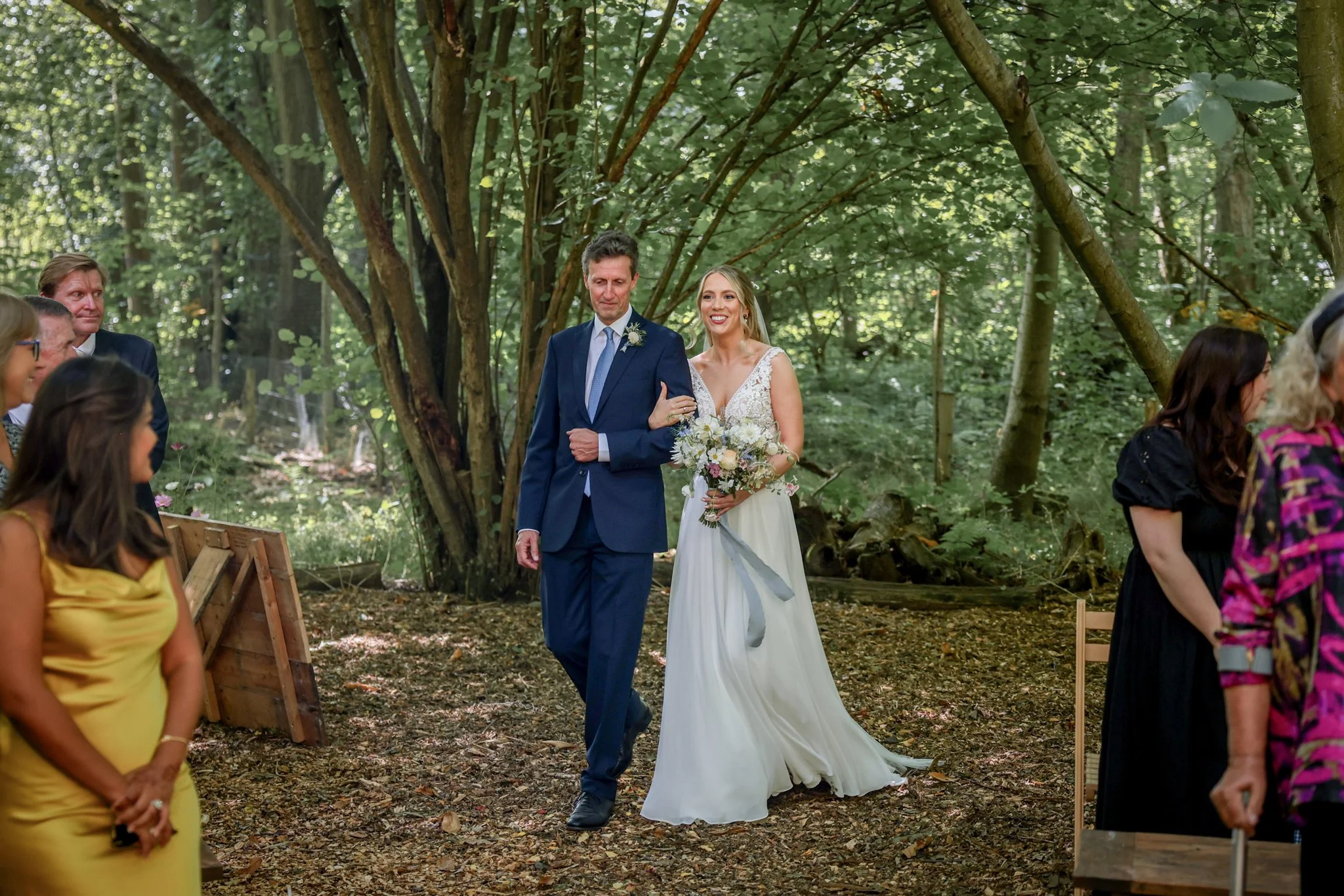 A wedding ceremony taking place outdoors in a forest with a bride and groom walking down the aisle. The bride is holding a bouquet and wearing a white dress, while the groom is in a dark suit. Several guests are seated along the aisle, surrounded by 