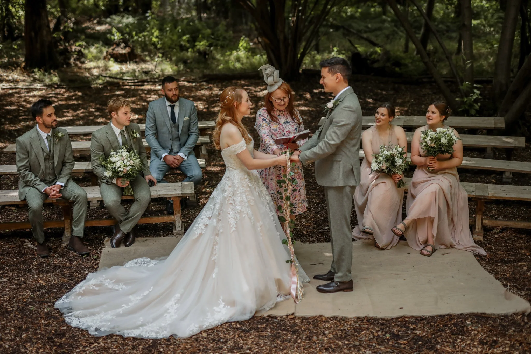 A bride and groom exchange vows during an outdoor wedding ceremony in a wooded area, with bridesmaids and groomsmen seated nearby.