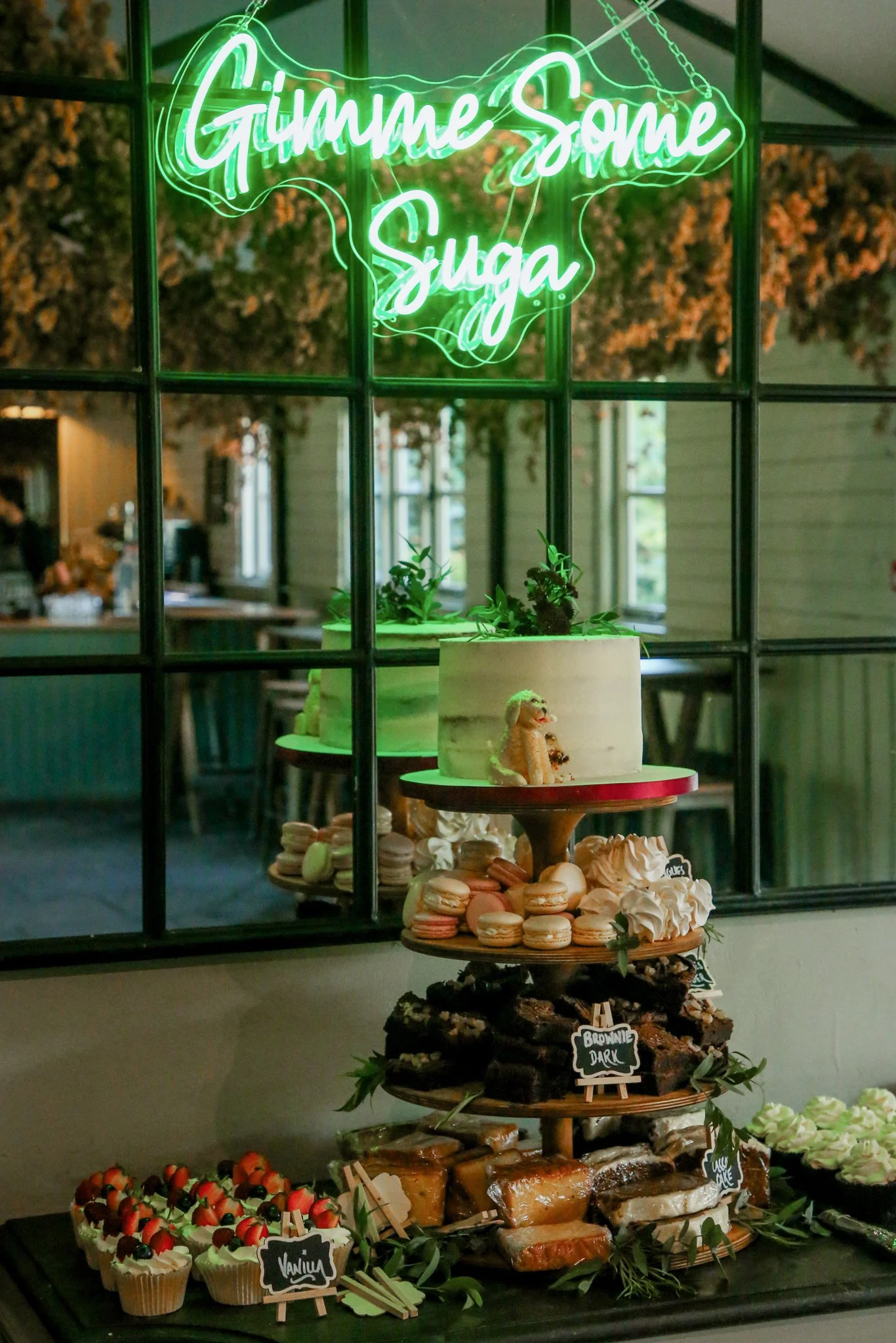 A dessert display with a neon sign reading 'Gimme Some Sugar' in green, featuring cupcakes, macarons, and various baked goods, set inside a room with windows and table.