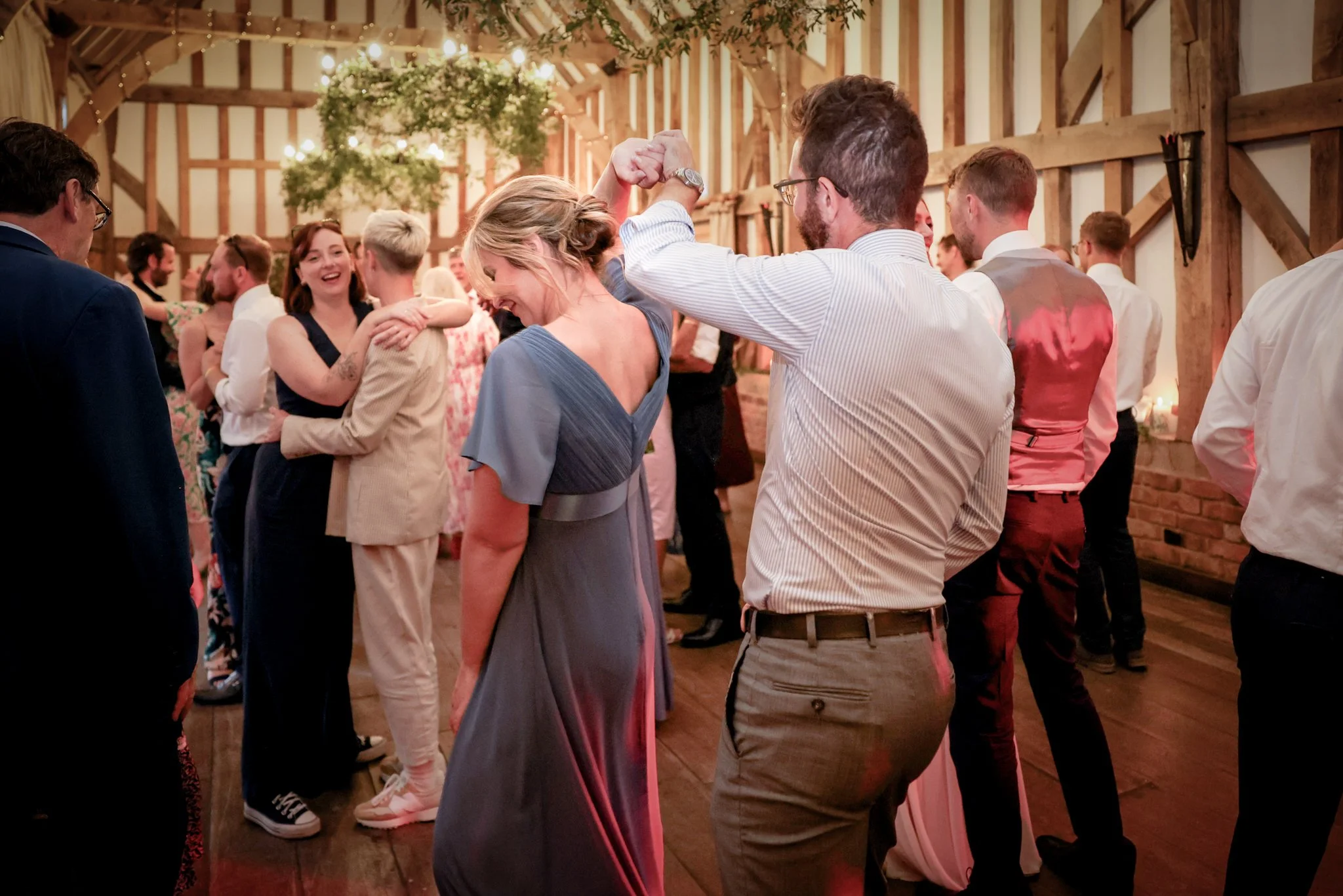 People dancing and celebrating at a wedding reception in a rustic barn with wooden beams, string lights, and decorations.