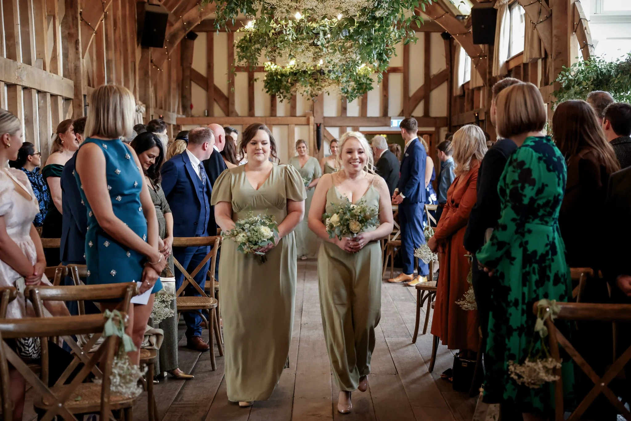 Wedding ceremony inside a rustic barn with guests seated on wooden chairs, two women in matching beige dresses walking down the aisle holding bouquets, ceiling decorated with greenery and string lights.