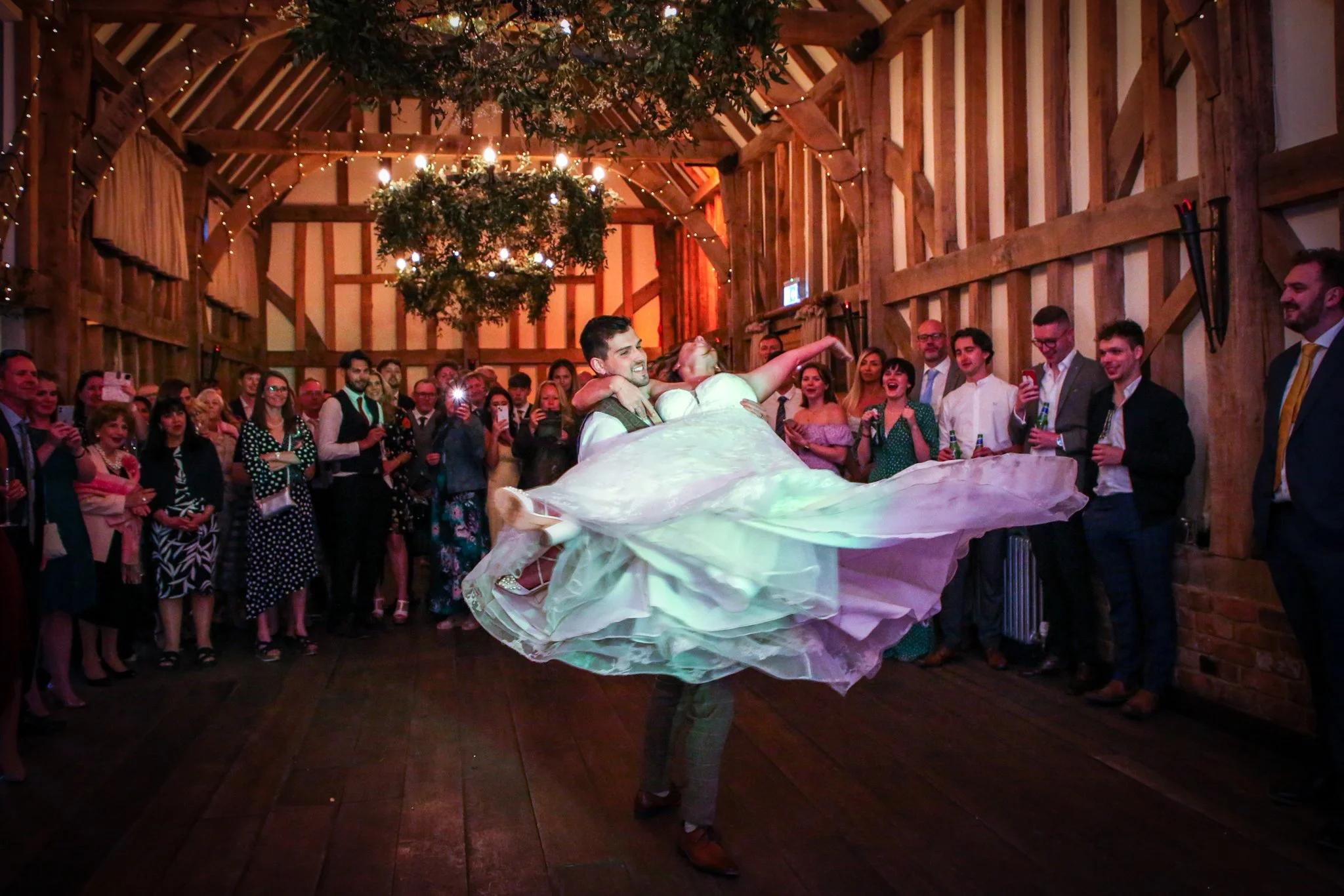 A bride and groom sharing a dance inside a rustic barn decorated with hanging greenery and string lights, surrounded by wedding guests.