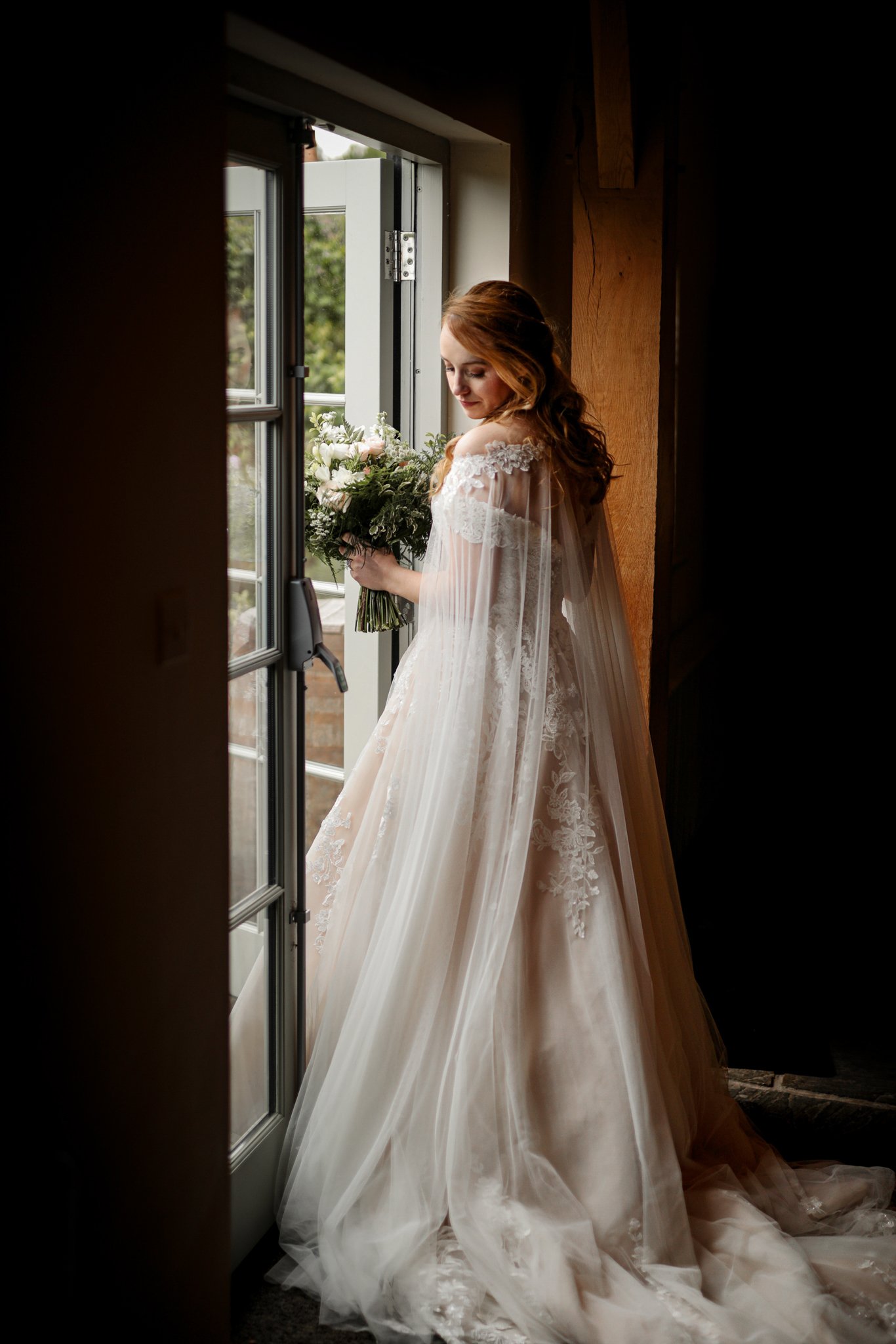 A bride with red hair in a wedding gown holding a bouquet of white and pink flowers near an open window in a wooden room.