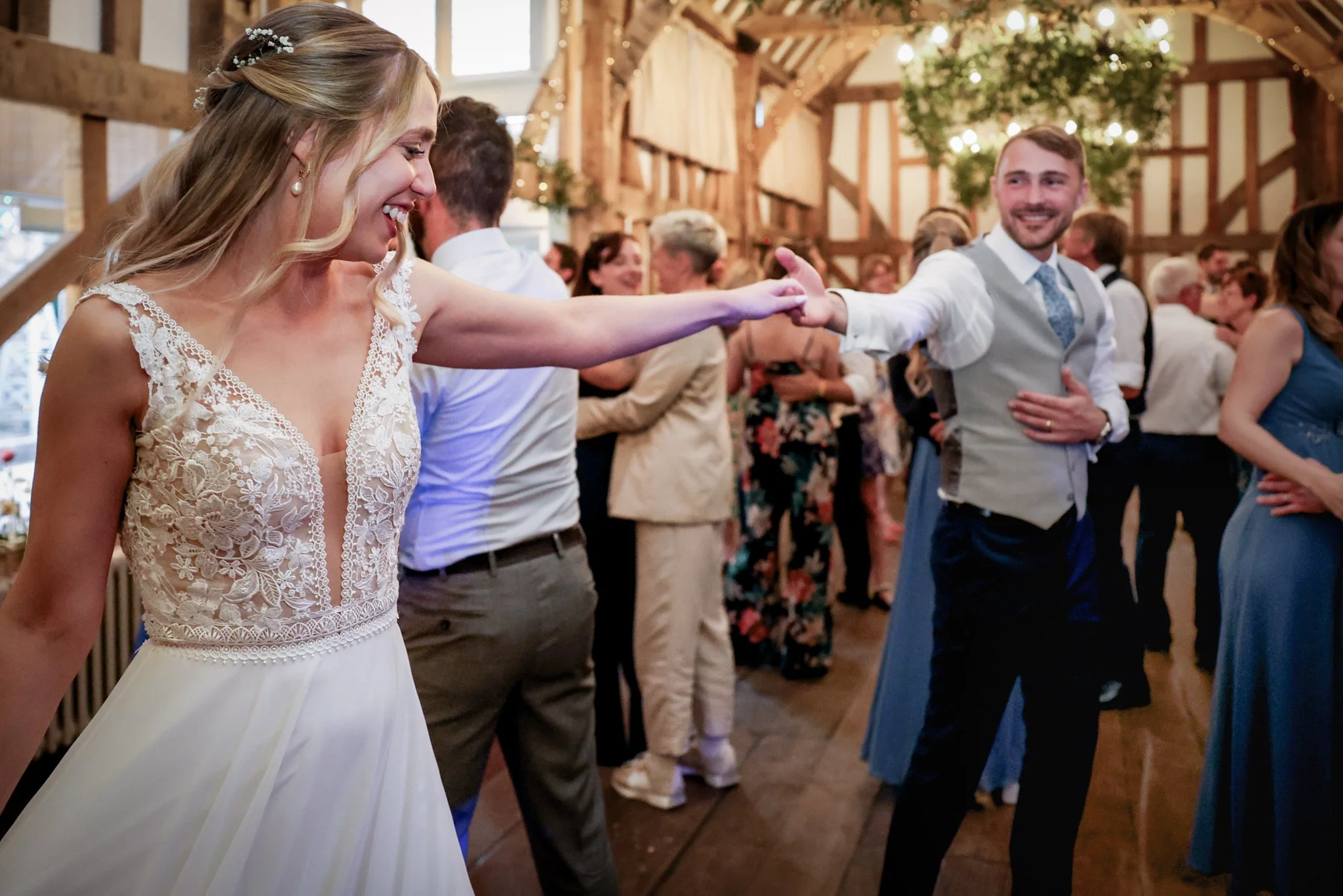 Bride and groom dancing at a wedding reception in a rustic barn with wooden beams and hanging lights, surrounded by guests.