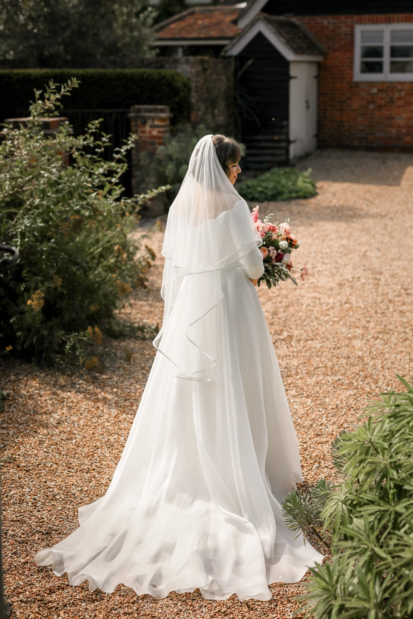 A bride stands outdoors in a garden, wearing a white wedding dress with a flowing train and a veil, holding a bouquet of pink and white flowers.