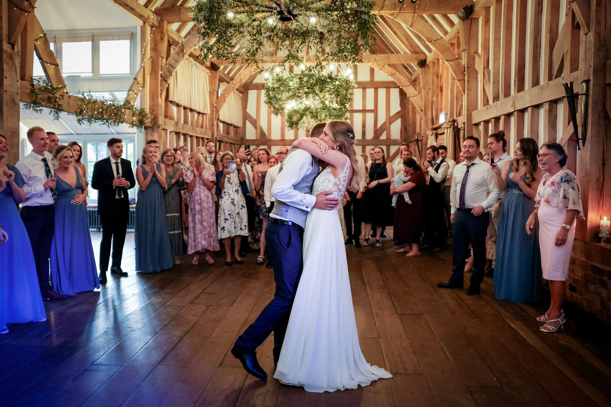 A couple dressed in wedding attire sharing a dance in a rustic barn during their wedding reception, surrounded by guests clapping and taking photos.