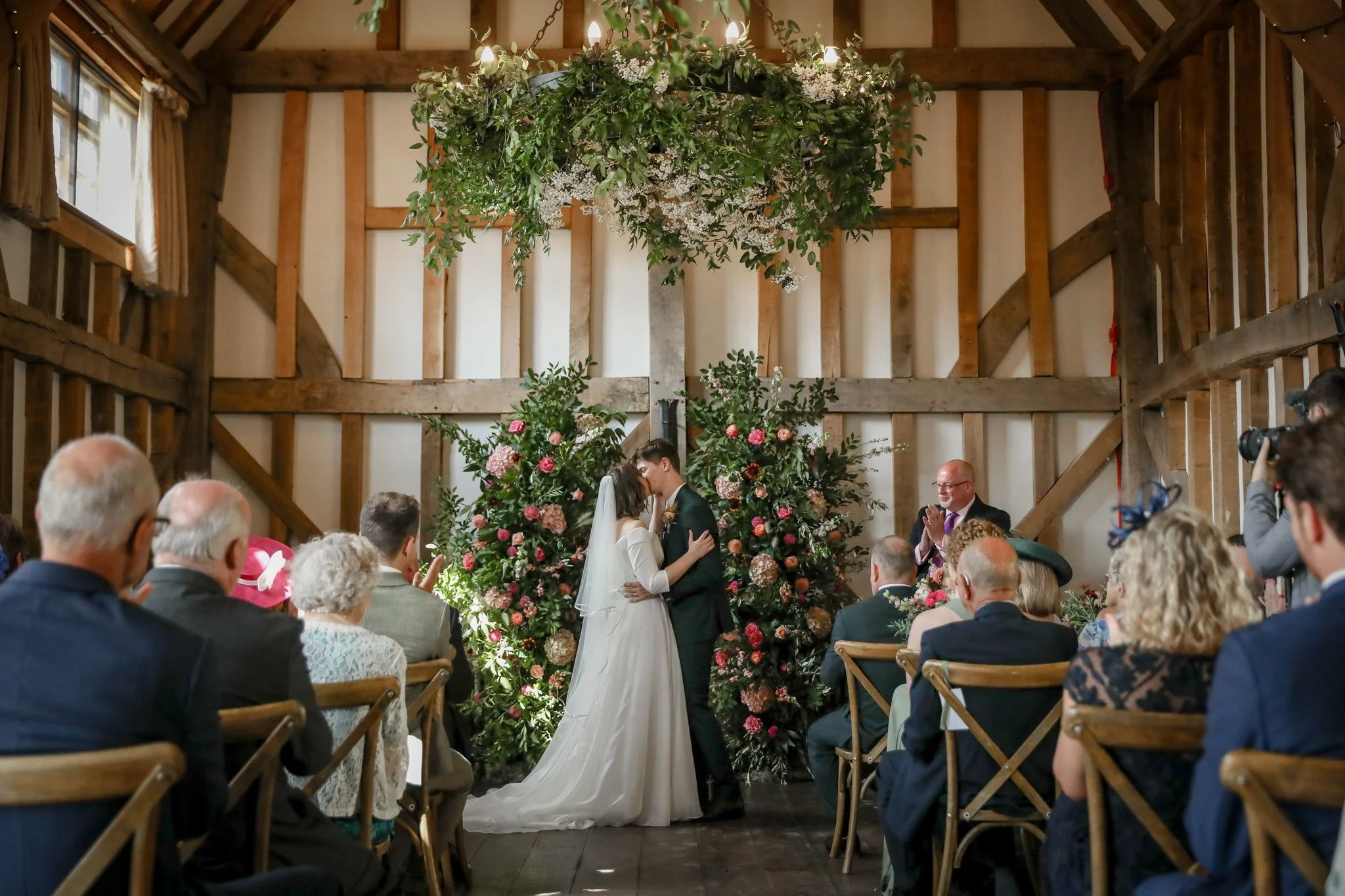 A wedding ceremony with a bride and groom kissing in front of a floral arch, surrounded by seated guests in a rustic barn setting.