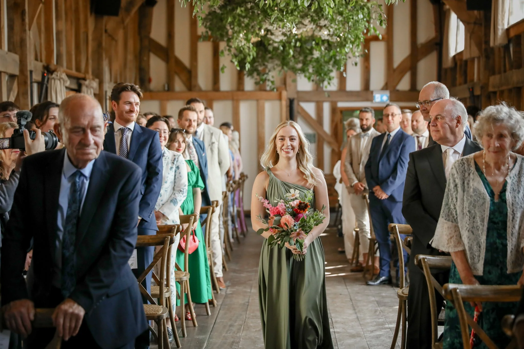A woman in a green dress holding a bouquet of flowers walking down the aisle at a wedding ceremony, surrounded by seated guests in a rustic barn setting.