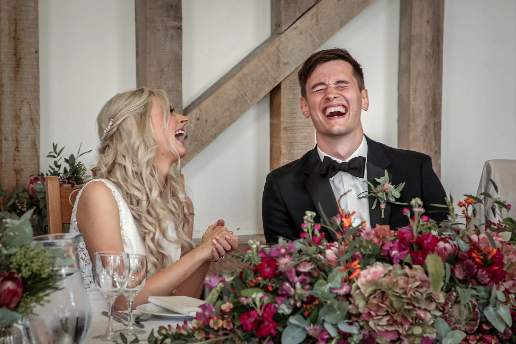 A bride and groom are laughing and holding hands at a wedding reception. The groom is wearing a black tuxedo with a bow tie, and the bride has long blonde hair and is wearing a white dress. There are colorful flowers on the table in front of them and