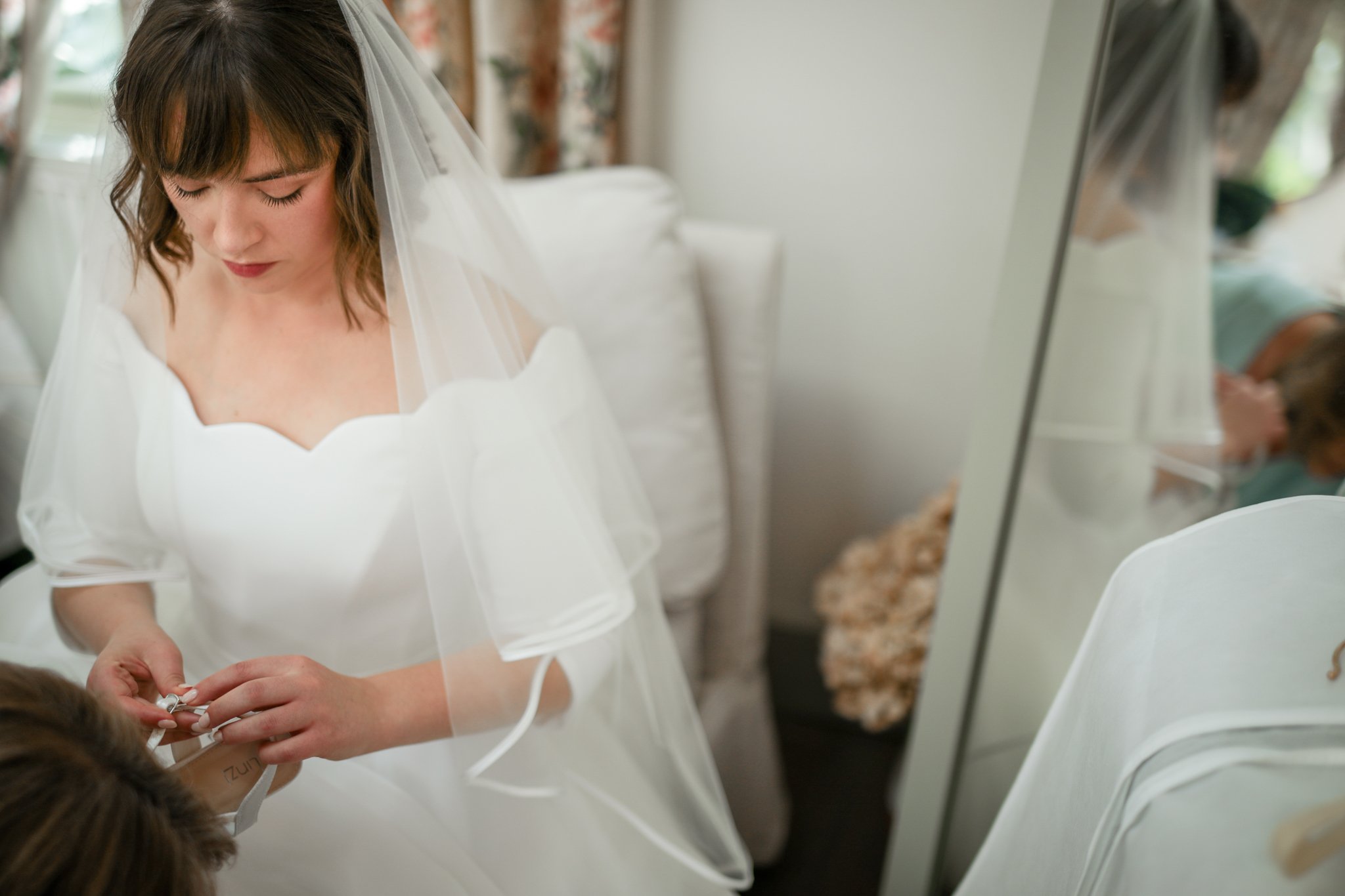 A woman dressed in white, possibly a bride, is adjusting jewelry while standing in front of a mirror. She has shoulder-length brown hair with soft waves and a sheer veil. The setting appears to be a room with floral curtains and a white chair, with a
