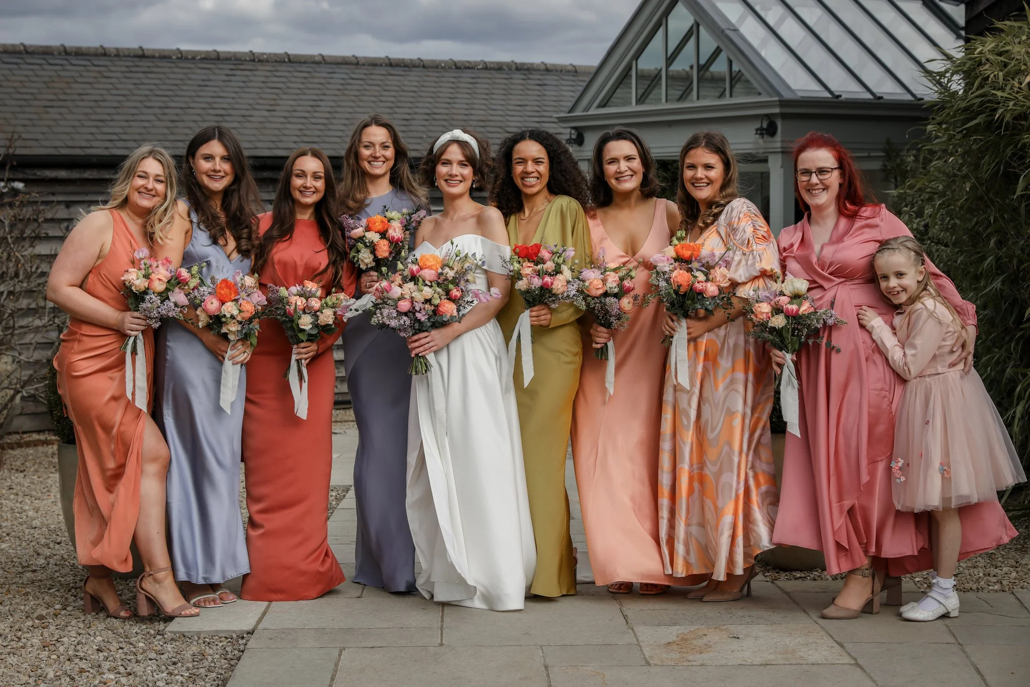 A group of ten women and one young girl celebrating a wedding outdoors on a cloudy day, all dressed in pastel and colorful dresses, holding bouquets of flowers, standing in front of a house with a slanted roof and a glass conservatory.