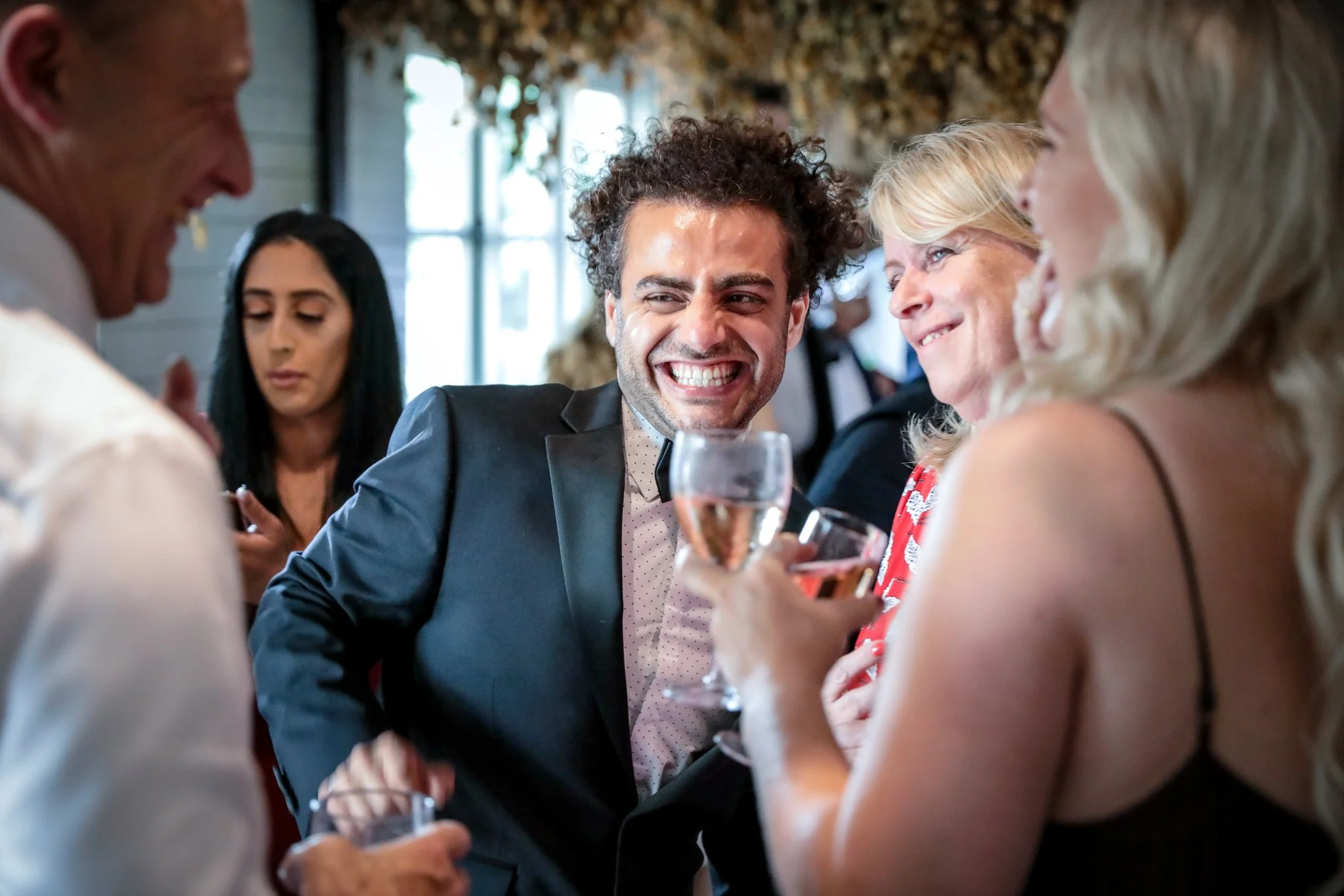 People celebrating, with a man in a suit smiling and holding a glass of champagne, surrounded by women in a social gathering.