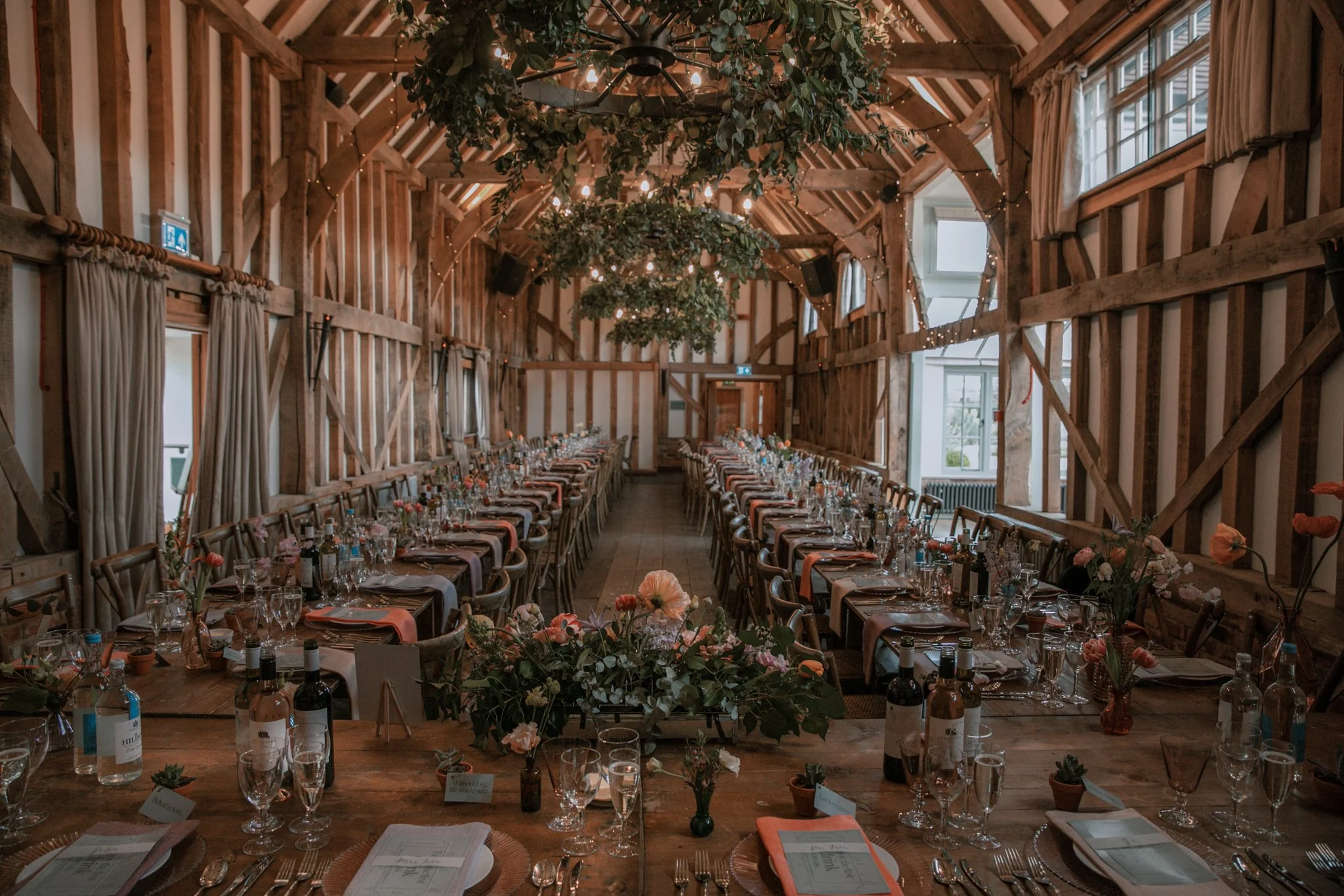Long banquet table set for a celebration inside a rustic wooden barn, decorated with flowers, wine bottles, and tableware.