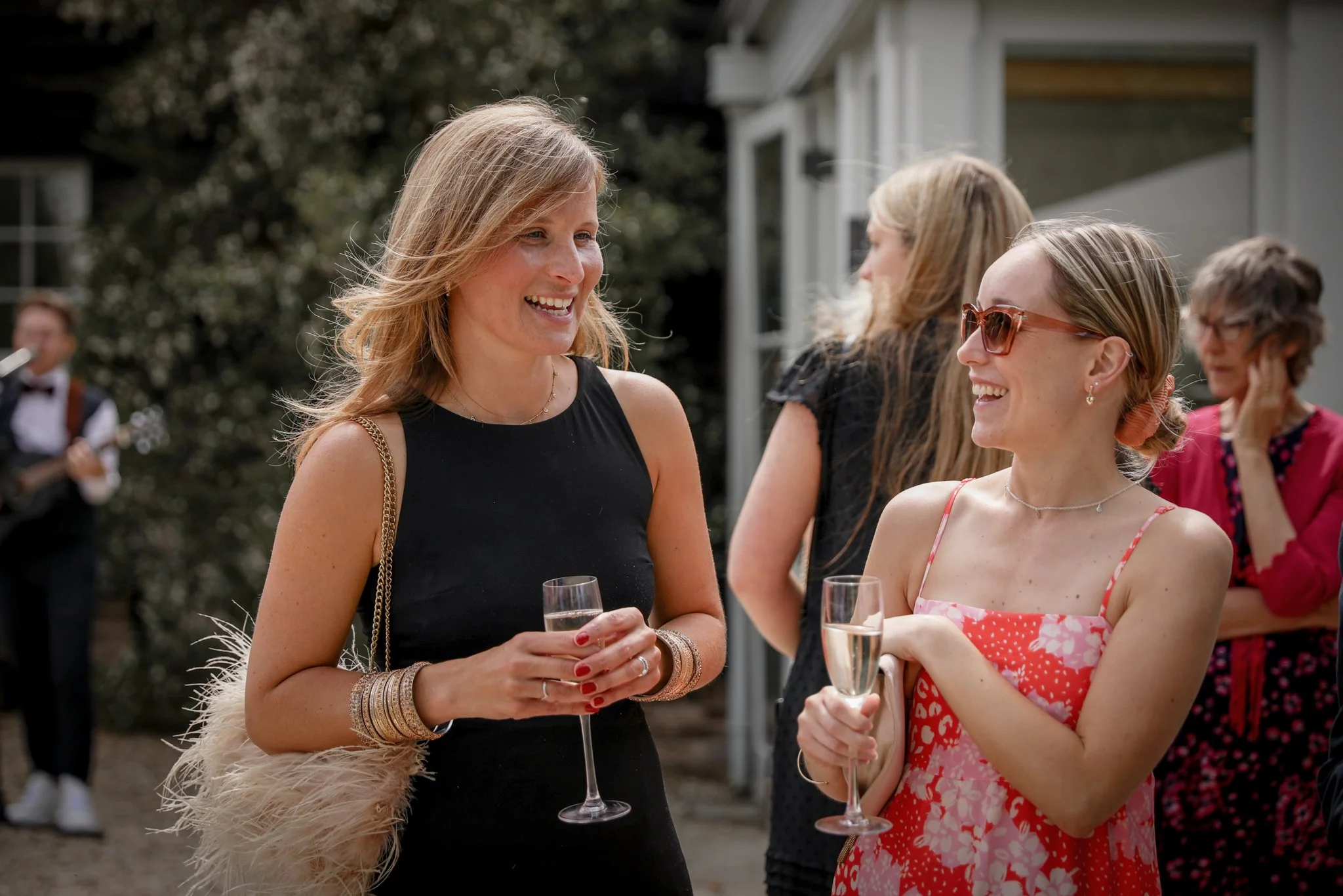 Two women are smiling and talking, holding glasses of champagne at an outdoor event. One woman has blonde hair, sunglasses, and is wearing a pink and white floral dress. The other woman has blonde hair, is wearing a black dress, and has multiple bang