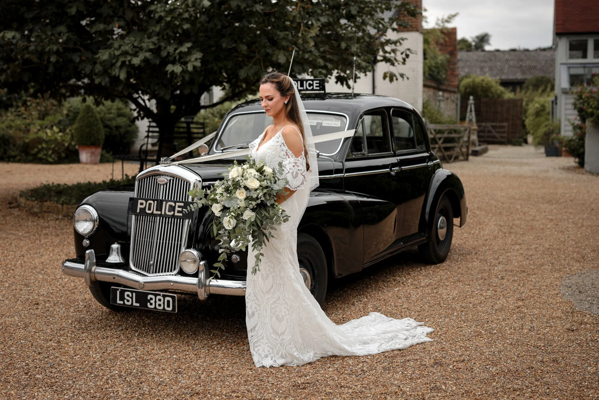 Bride in a white wedding dress holding a bouquet, standing beside a vintage black police car decorated for a wedding, parked on a gravel driveway with trees and houses in the background.