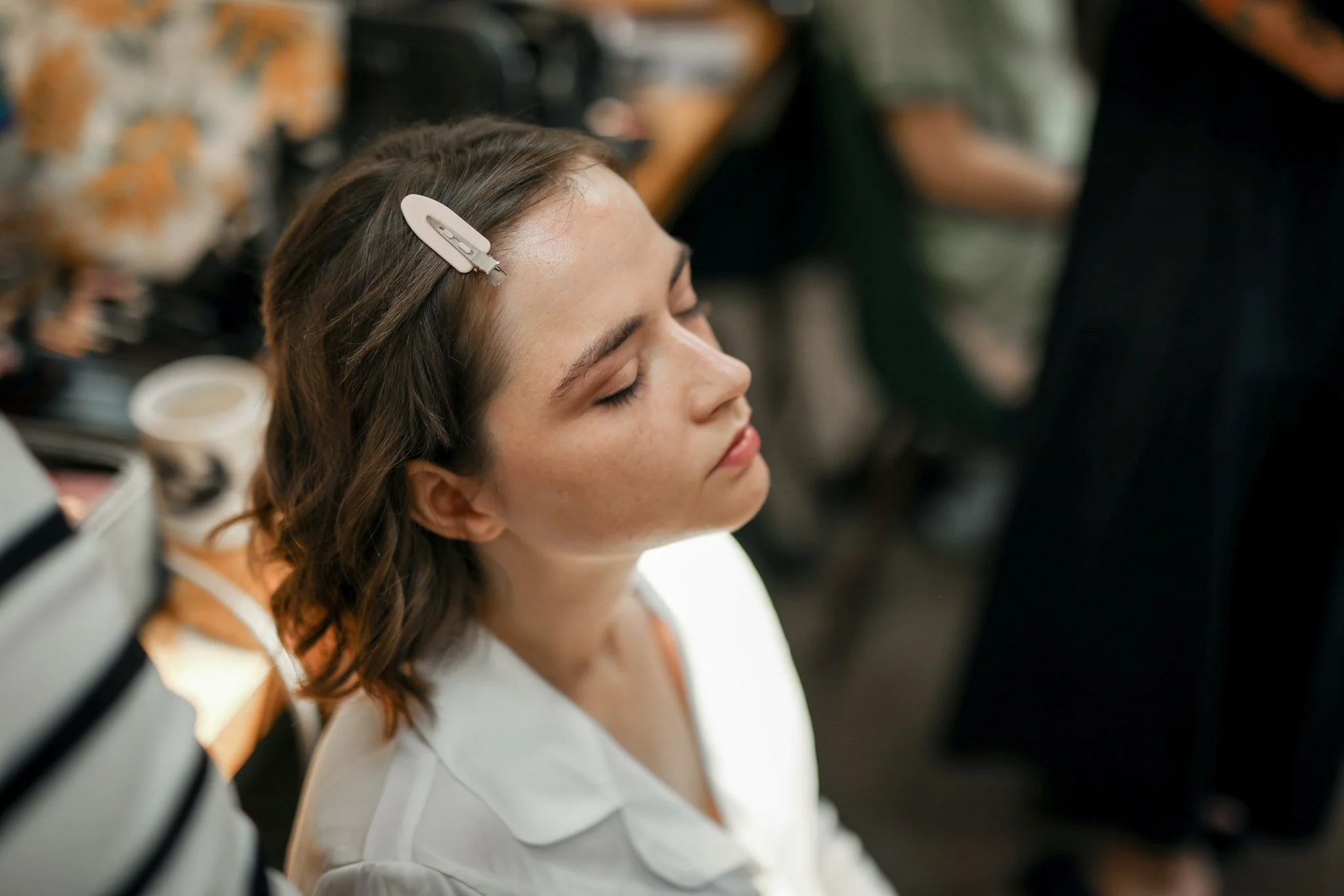 A young woman with brown hair, closed eyes, and a hair clip in her hair, lying down with her head resting on a surface, possibly during a makeup or beauty treatment.