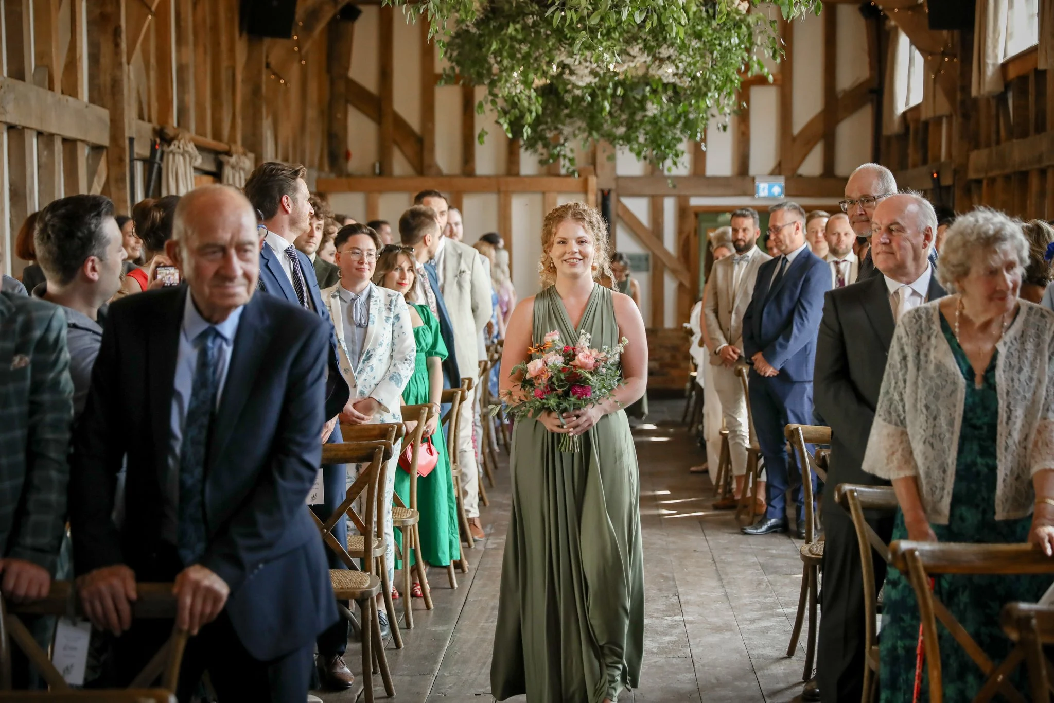 A woman in a green dress holding a bouquet of flowers walking down the aisle during a wedding ceremony in a rustic barn with wooden beams, surrounded by seated guests.