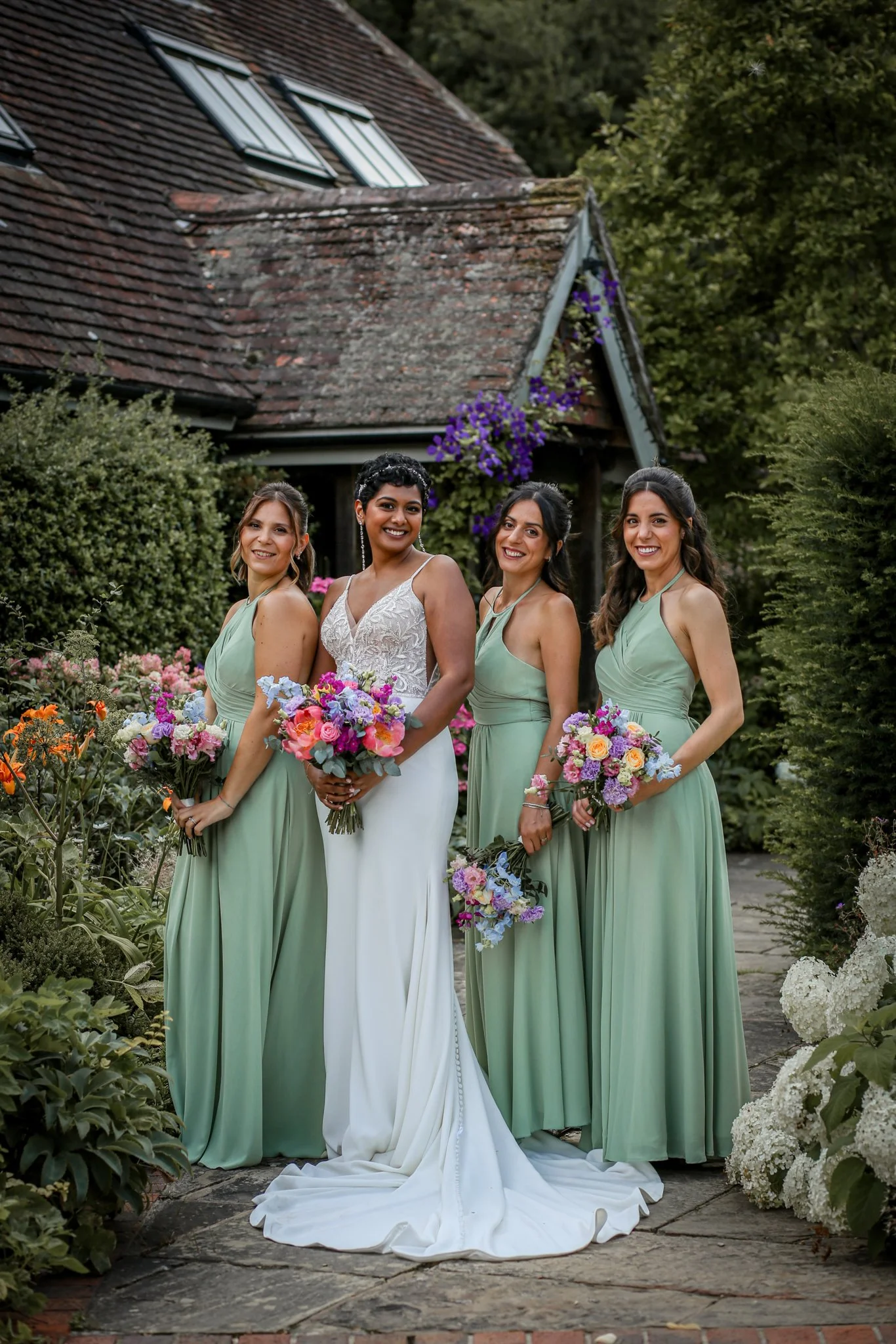 Four women in a garden: one bride in a white wedding dress, three bridesmaids in matching green dresses, holding floral bouquets, standing on stone path, greenery and flowers around, old house with tiled roof in background.