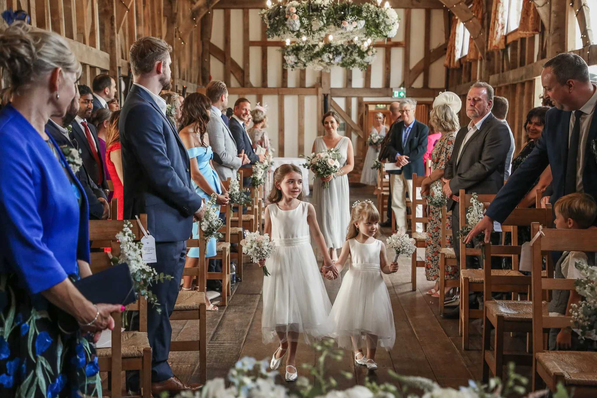 A wedding ceremony inside a rustic wooden barn with floral decorations. Two young girls in white dresses walk down the aisle holding flowers, with the bride and guests visible on either side.