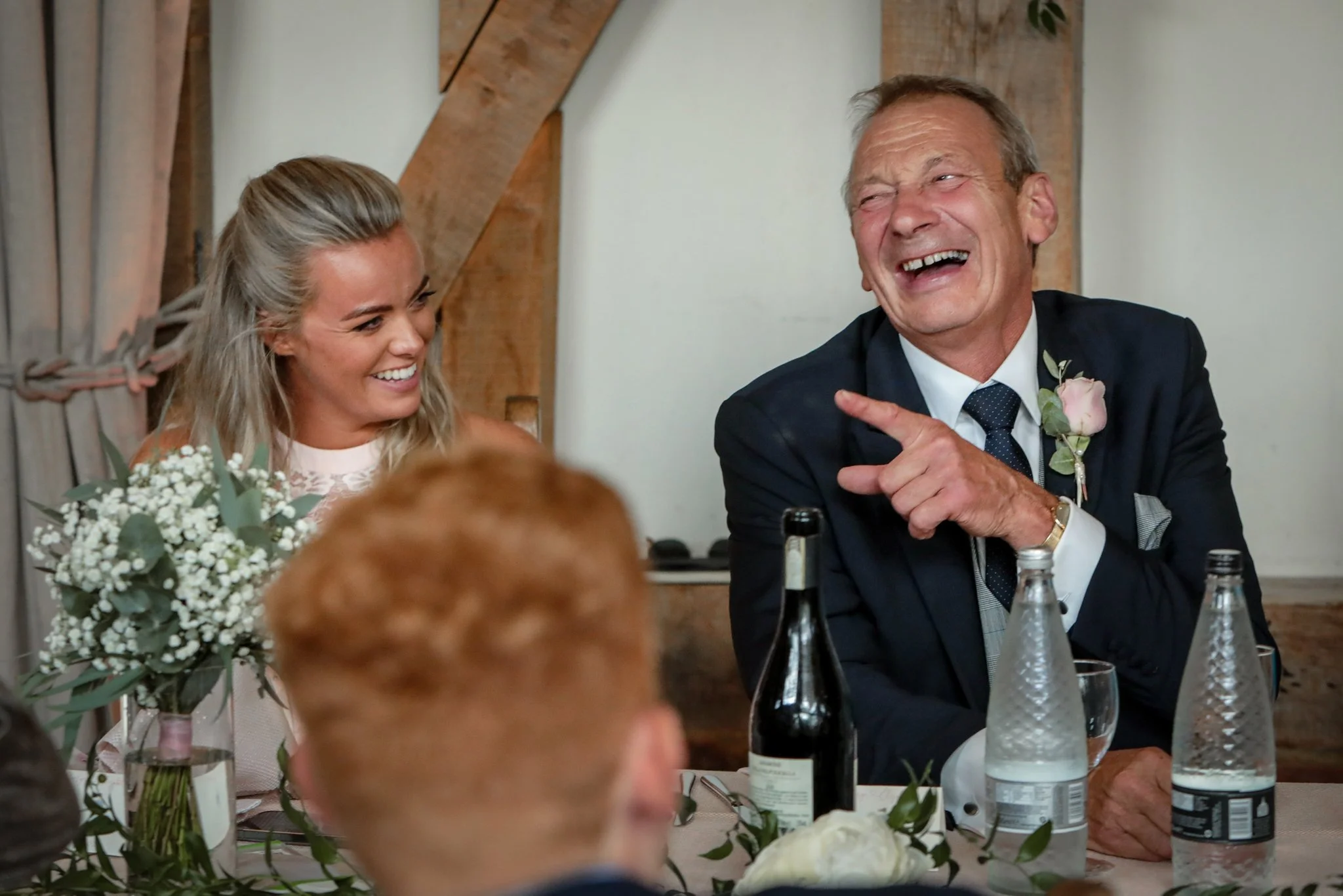 A happy bride and an older man laughing at a wedding reception, sitting at a table with floral arrangements and bottles of beverages.