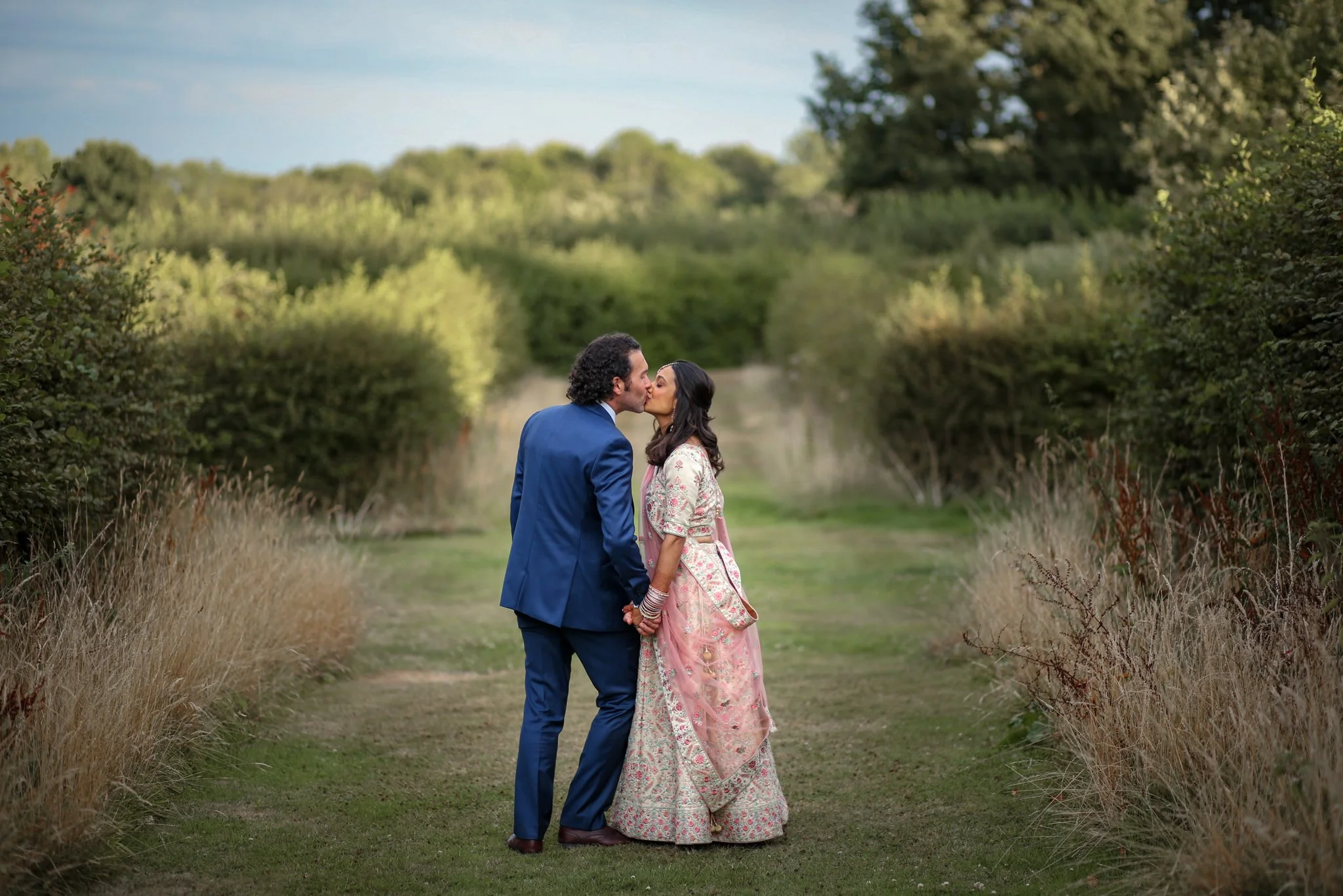 A couple is kissing outdoors on a grassy path, surrounded by tall bushes and trees, with a clear blue sky overhead.