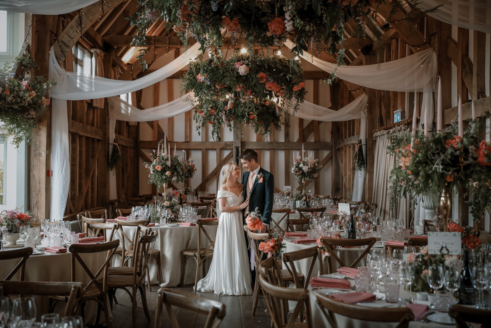 A bride and groom standing together in a rustic wedding reception hall decorated with hanging greenery, flowers, and white drapes, with tables set for a celebration.
