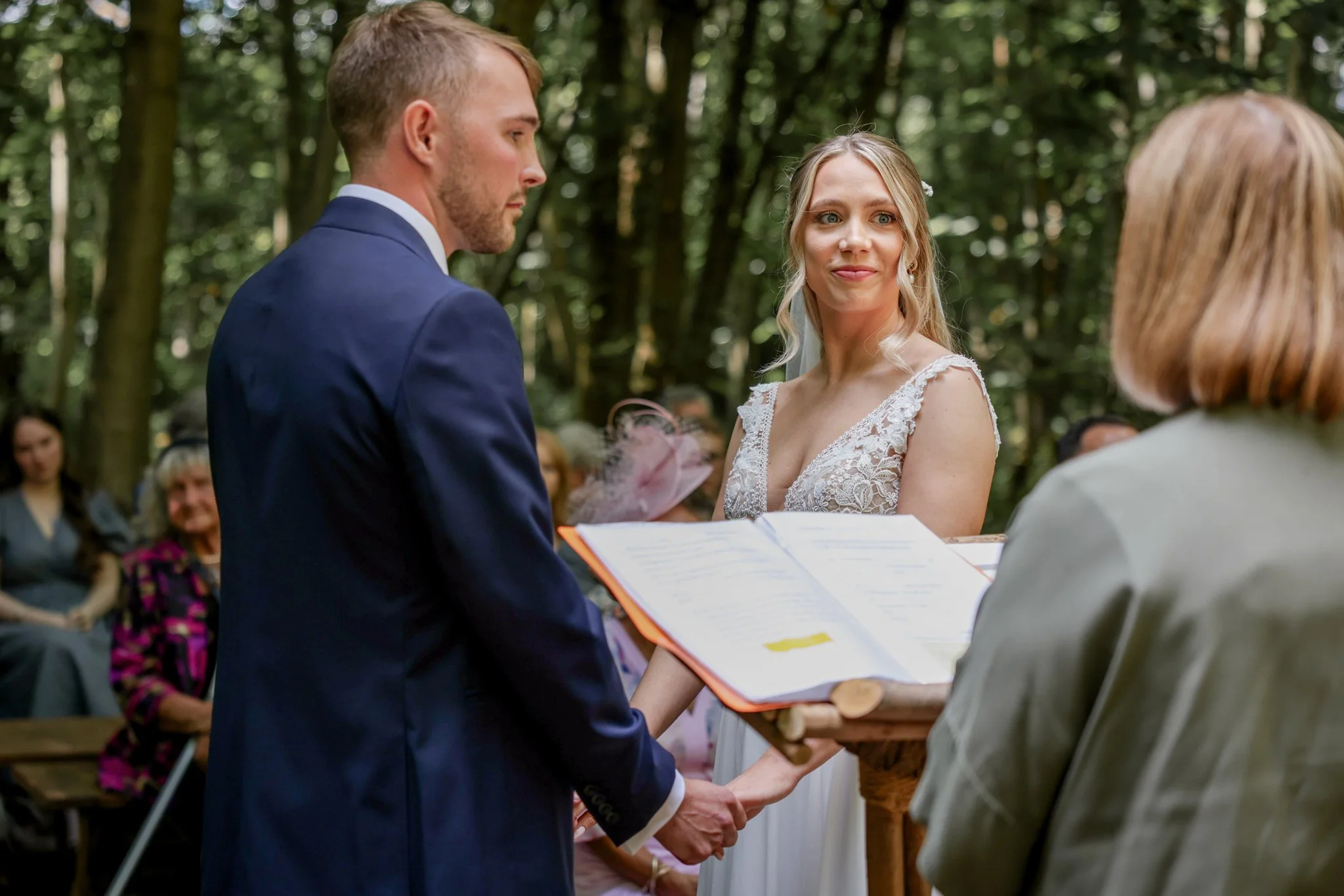 A wedding ceremony in a forest with a groom and bride holding hands, exchanging vows, with an officiant reading from a book, and guests in the background.