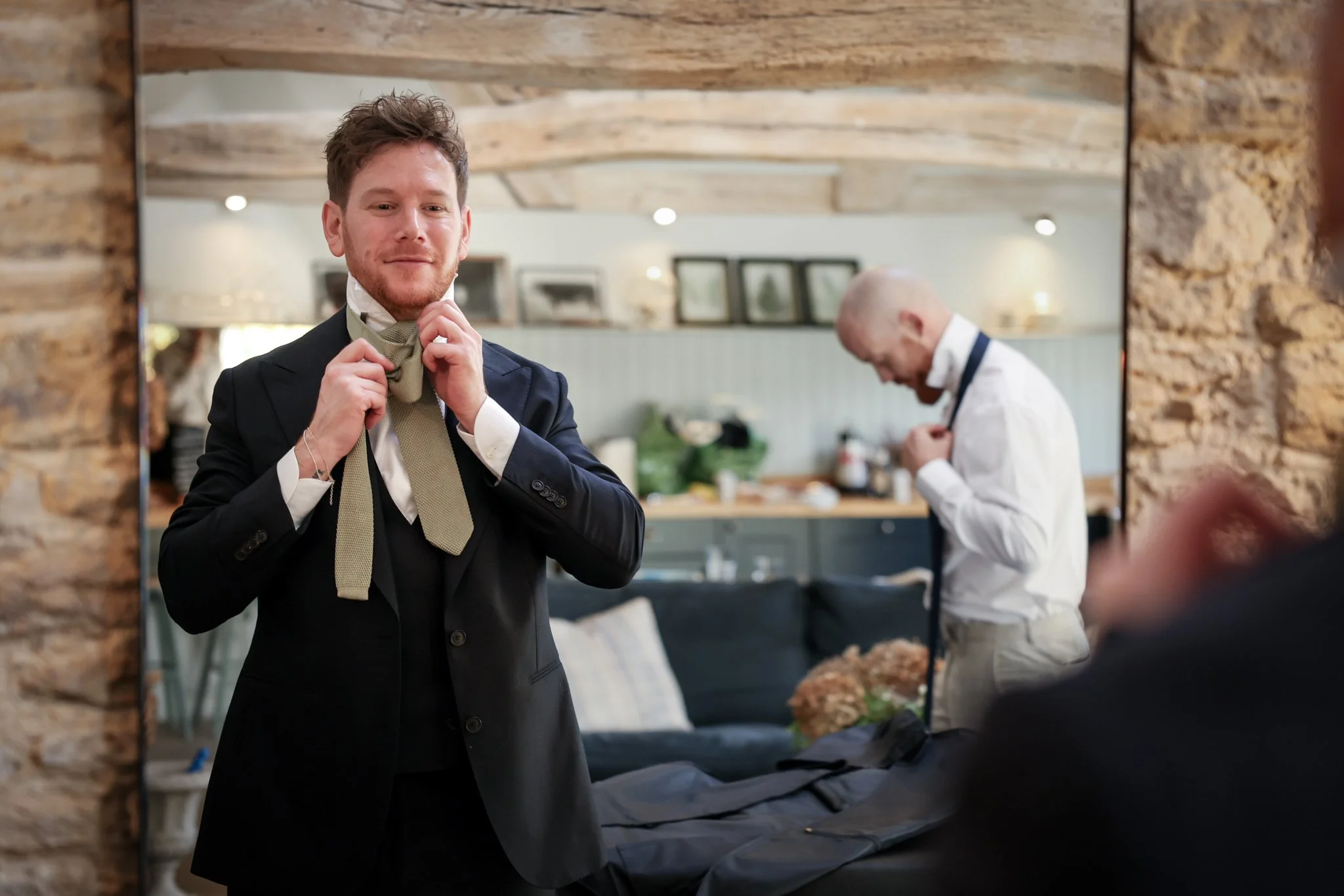 A young man in a suit is adjusting his tie while looking at himself in a mirror. Another man is in the background, wearing a white shirt and apron, preparing food in a kitchen with stone walls.