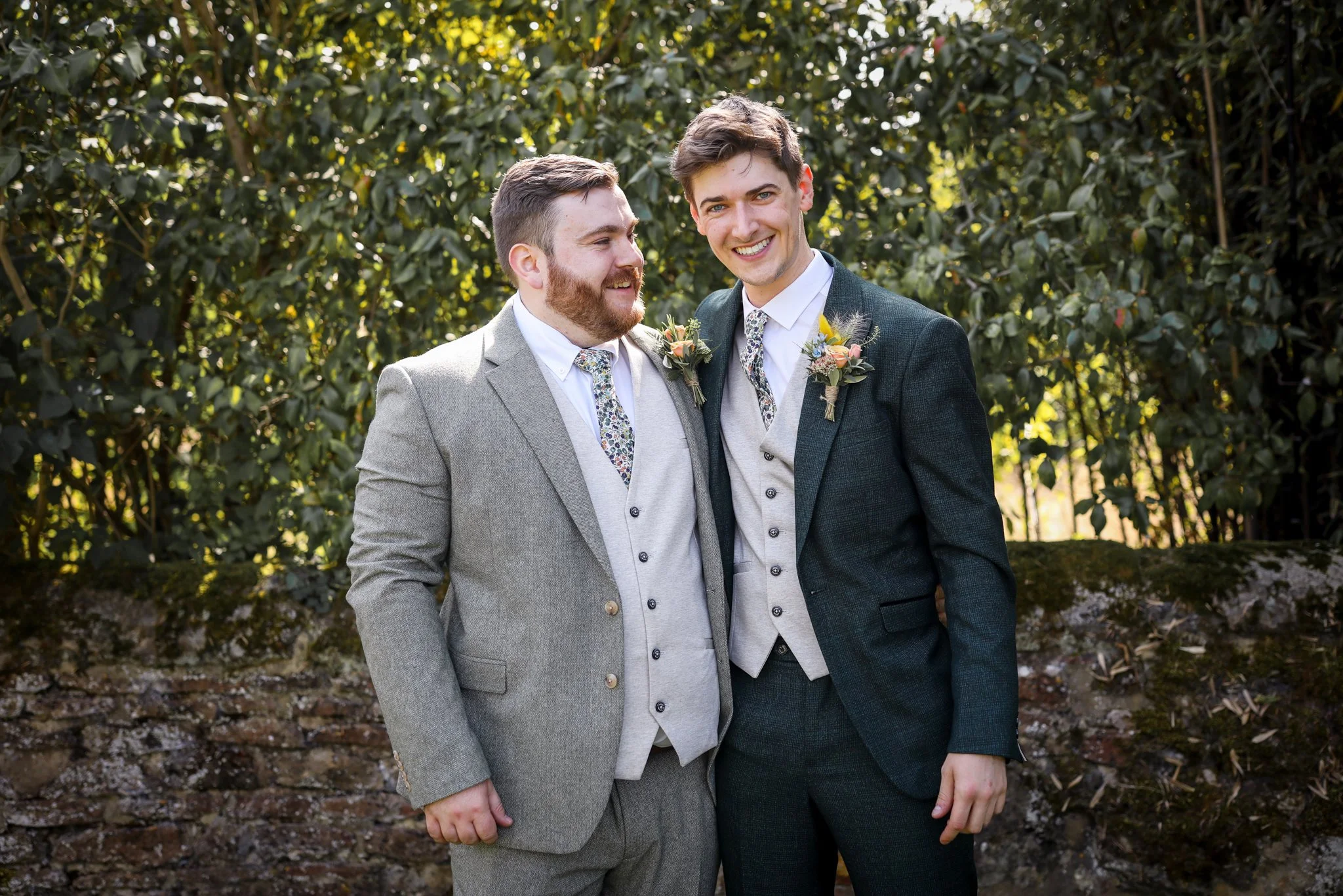 Two men in suits, one with a beard and the other clean-shaven, standing outdoors in front of green foliage, smiling and holding hands.