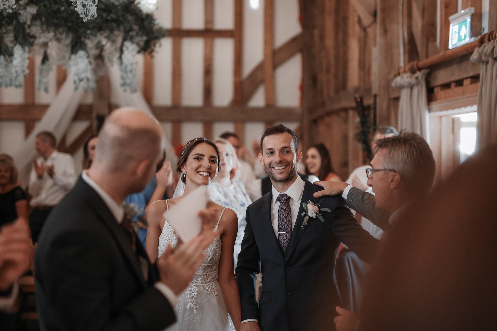 A bride and groom smiling at each other during a wedding reception inside a rustic barn with exposed wooden beams and walls, surrounded by guests.
