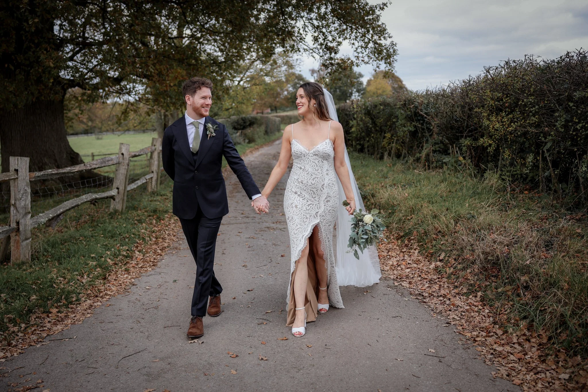 A newlywed couple walking hand in hand on a rural path, smiling at each other, with trees and bushes on either side and a cloudy sky overhead.