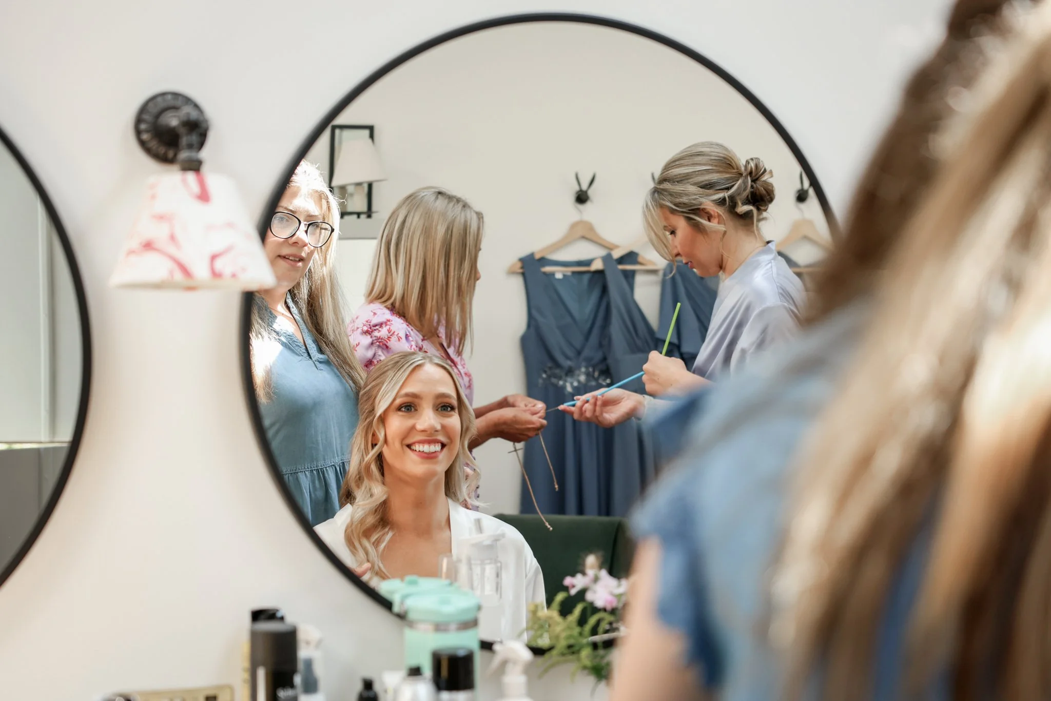 A woman with blonde hair and a white robe smiling in front of a mirror, with other women assisting her, possibly during her wedding preparations, in a room with hanging dresses and decorated with flowers.
