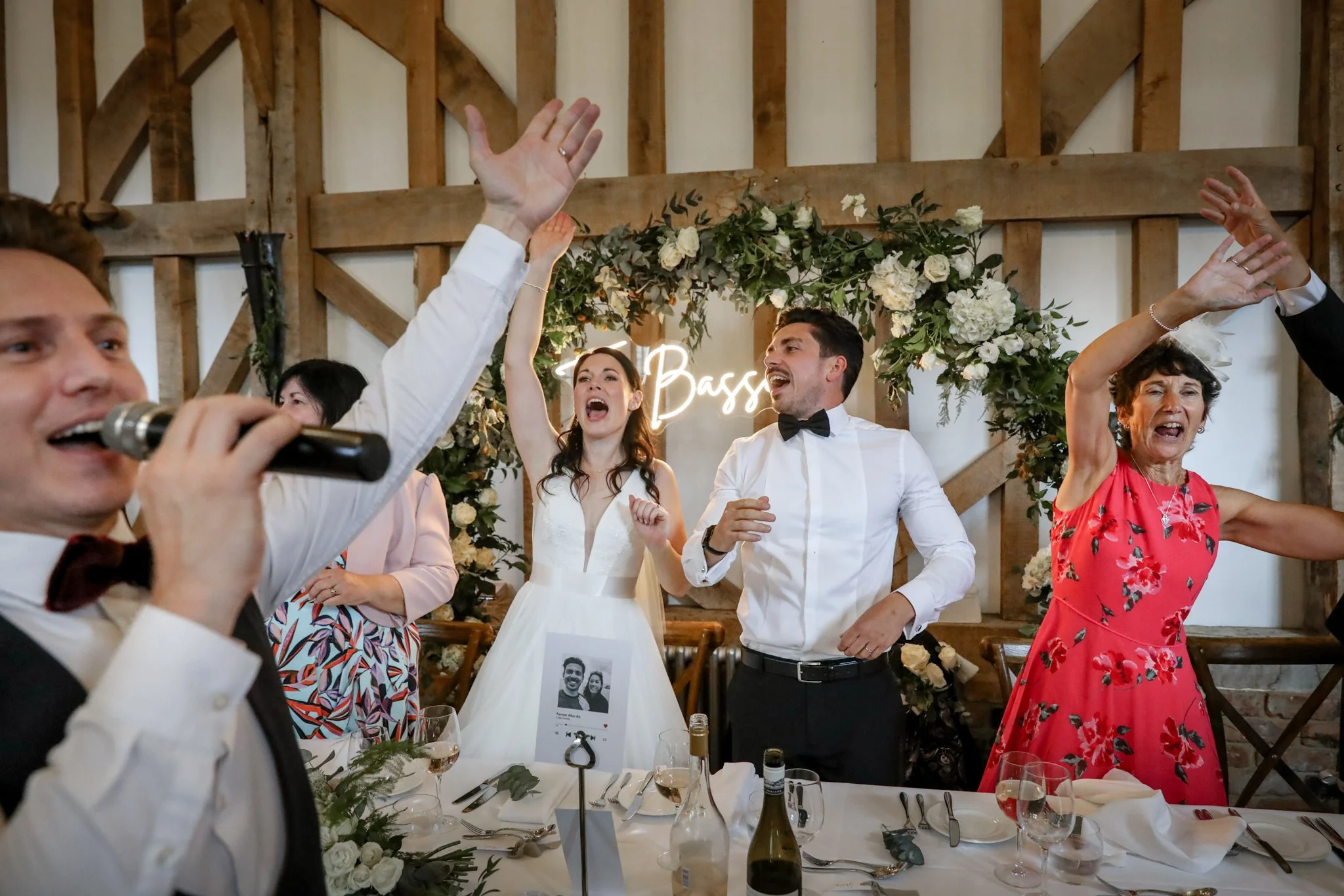 People celebrating at a wedding reception, dancing and singing with a woman in a white dress and a man in a white shirt and black bow tie at the center, surrounded by women in colorful dresses. There are floral decorations and a lit sign in the backg