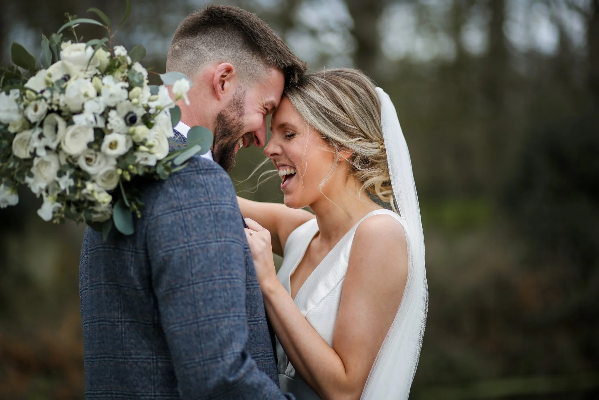 A happy couple, a man and a woman, sharing an intimate moment outdoors, with their foreheads touching and eyes closed. The man is holding a bouquet of white flowers. The background is blurred greenery.