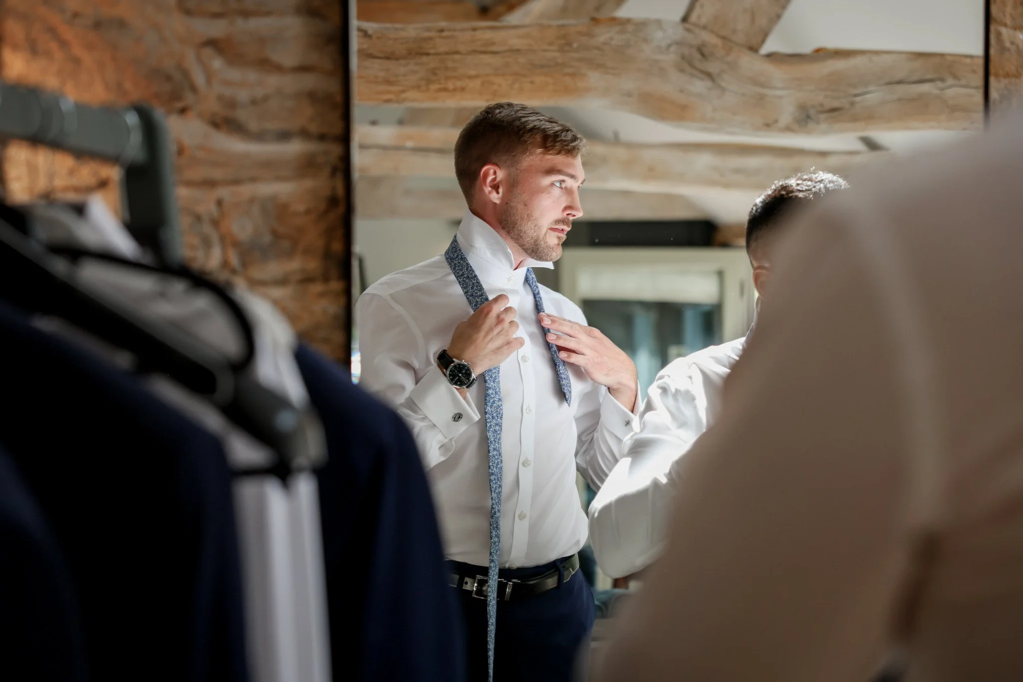 A man in a white shirt putting on a tie, looking into a mirror, in a room with wooden walls.
