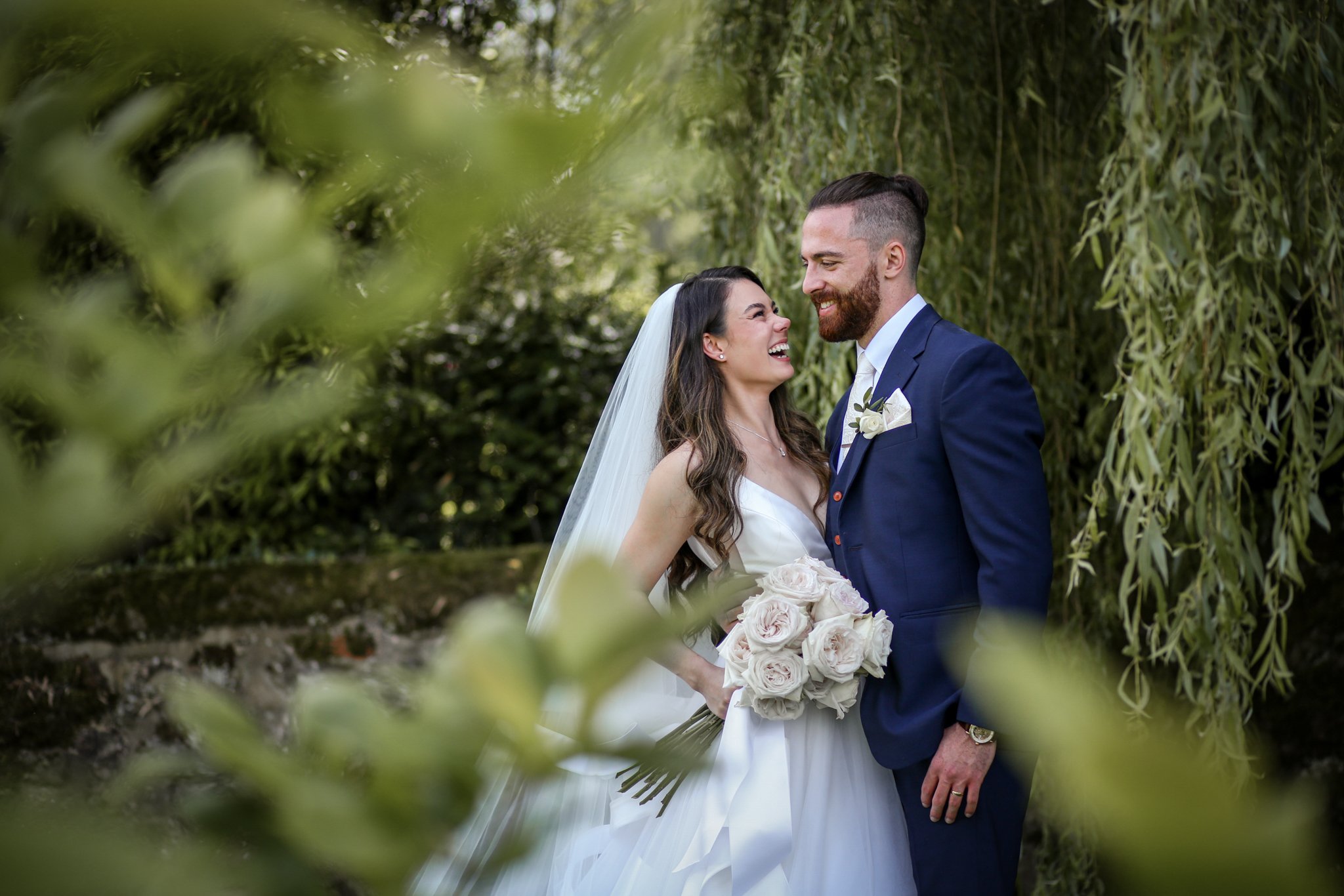 A bride and groom sharing a joyful moment outdoors, with greenery and hanging branches in the background. The bride is wearing a white wedding dress and veil, holding a bouquet of flowers, while the groom is dressed in a navy blue suit with a white s