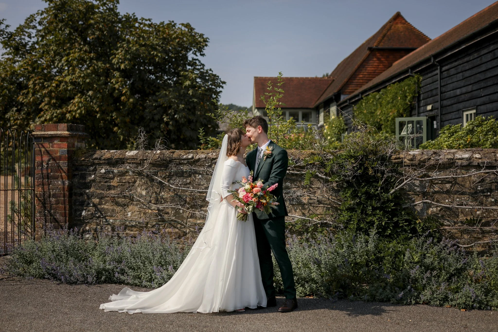 A bride and groom kiss outdoors on their wedding day, with the bride holding a bouquet of flowers. They stand in front of a rustic brick and wooden fence with greenery and a stone wall.