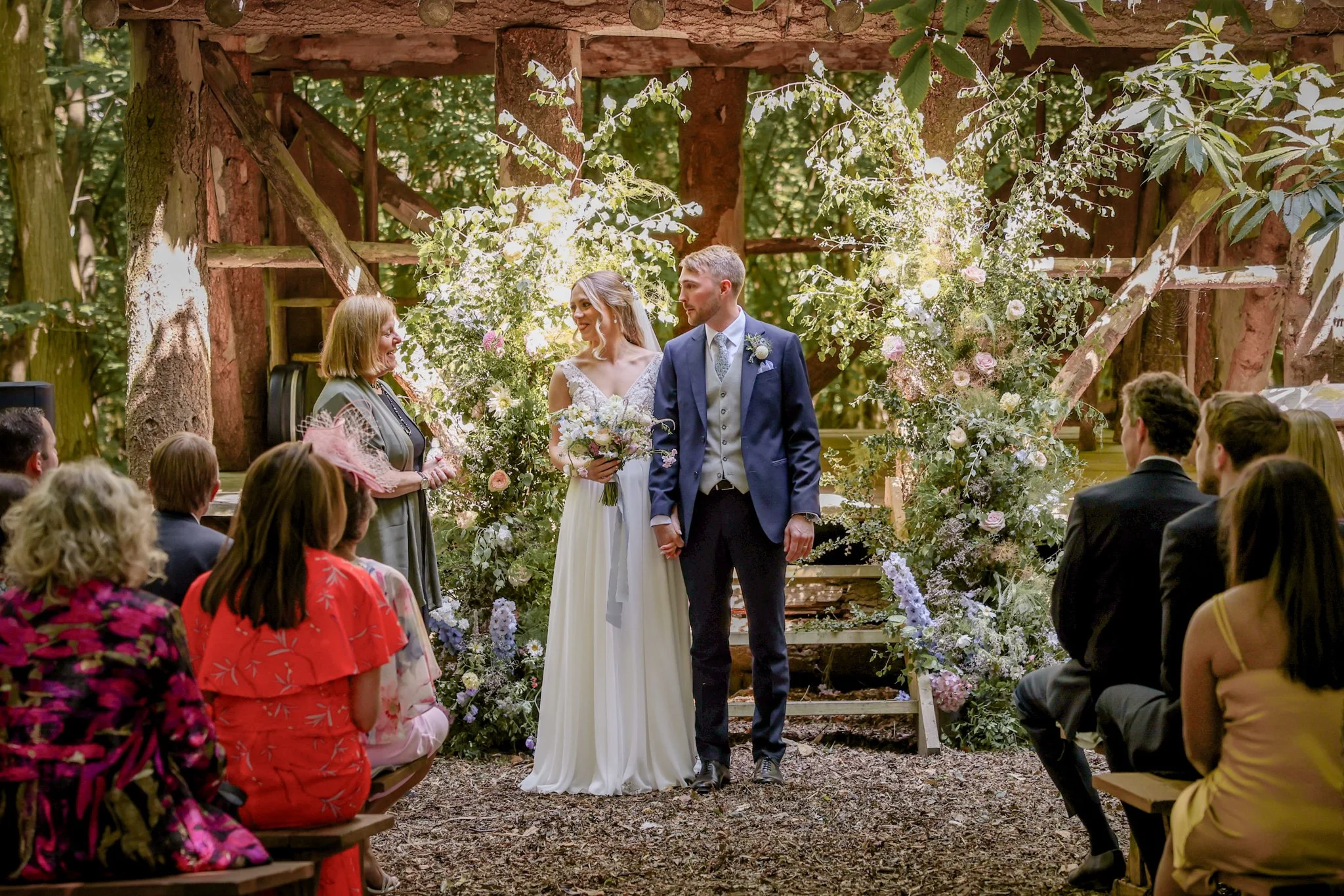 A wedding ceremony taking place outdoors in a forest, with a bride and groom standing at the altar decorated with flowers, surrounded by seated guests.