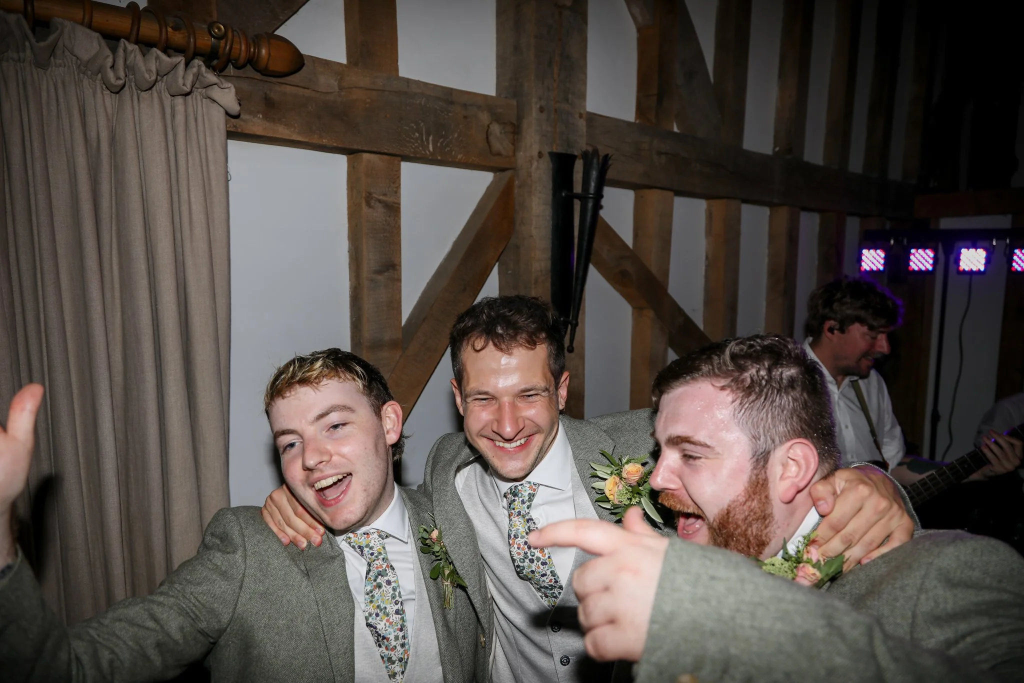 Group of men dressed in suits at a celebration, smiling and taking a selfie inside a rustic venue with wood beams and a curtain.