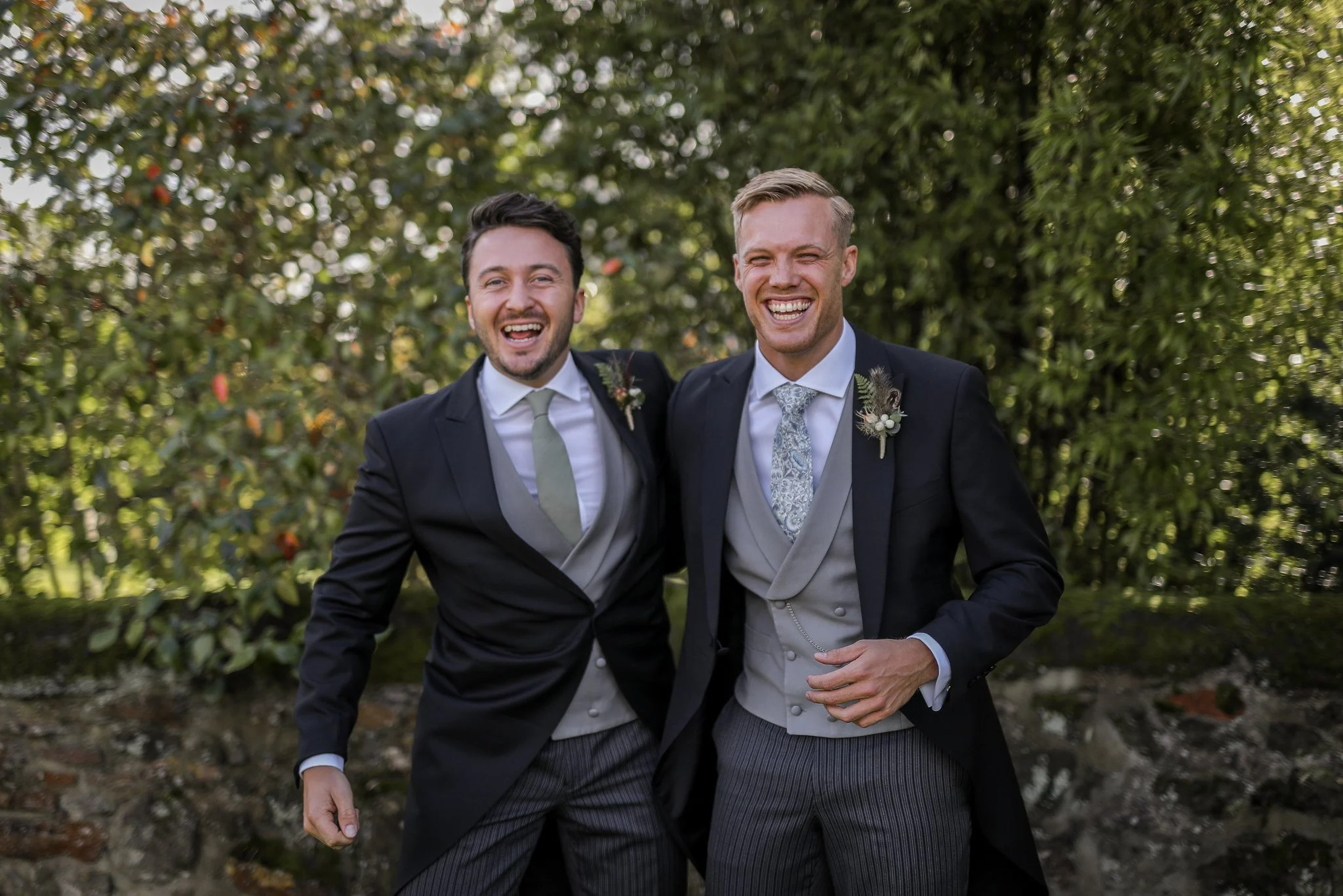 Two men in suits smiling and walking outdoors among greenery. One has a boutonniere with a feather and white flower on his lapel.