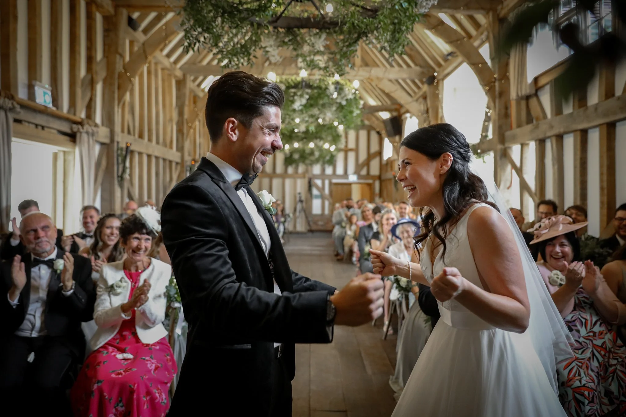 A bride and groom are smiling and holding hands during their wedding ceremony in a rustic barn with wooden beams and string lights, while guests clap and cheer.