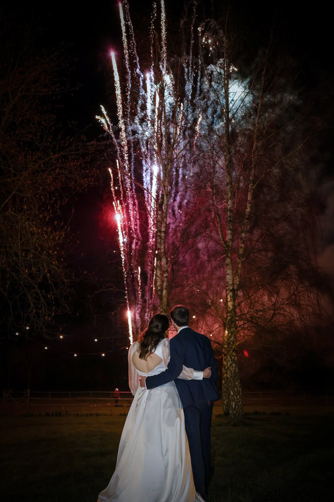 A bride and groom in wedding attire standing outdoors at night, watching fireworks light up the sky behind bare trees.