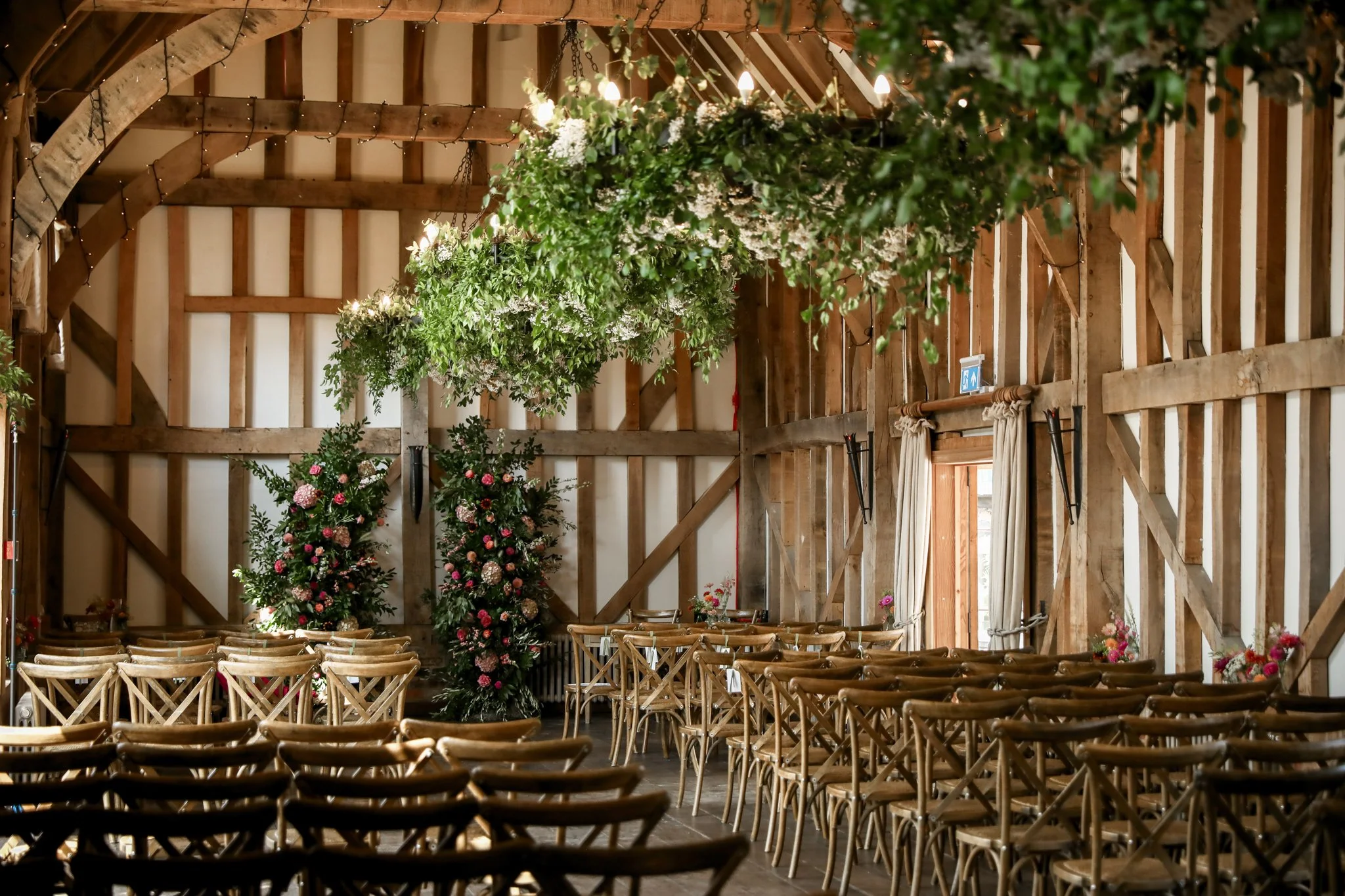 Indoor wedding or event space with wooden chairs arranged for guests, decorated with large floral arrangements of pink and white flowers, hanging greenery, and string lights on the wooden ceiling.