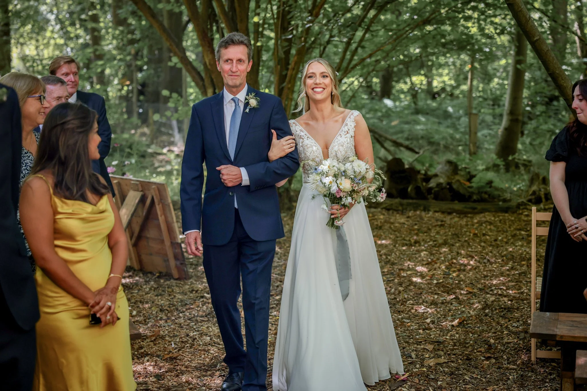 A bride walking down the aisle arm-in-arm with her father during a wedding ceremony in an outdoor wooded setting.