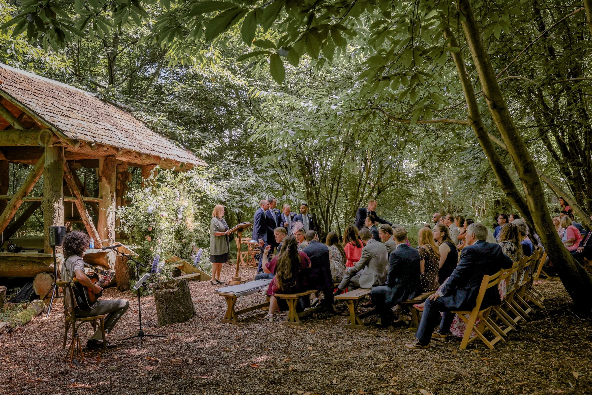 A gathering of people attending an outdoor event in a wooded area with a wooden structure and trees nearby. Some people are seated on wooden benches, while others are standing and listening to a woman speaking at a small podium. A musician is sitting