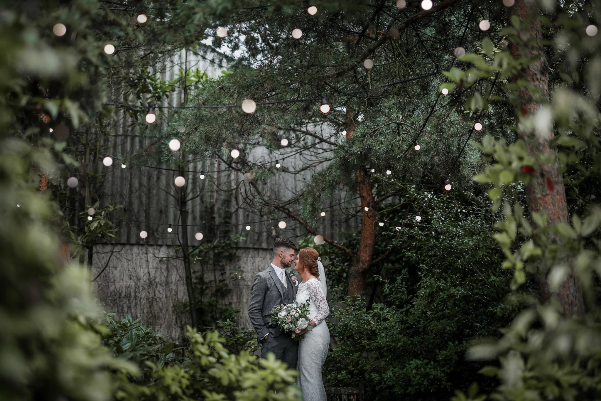 A bride and groom standing closely together outdoors, holding hands and looking at each other, under a canopy of string lights hanging from trees, with lush greenery surrounding them.