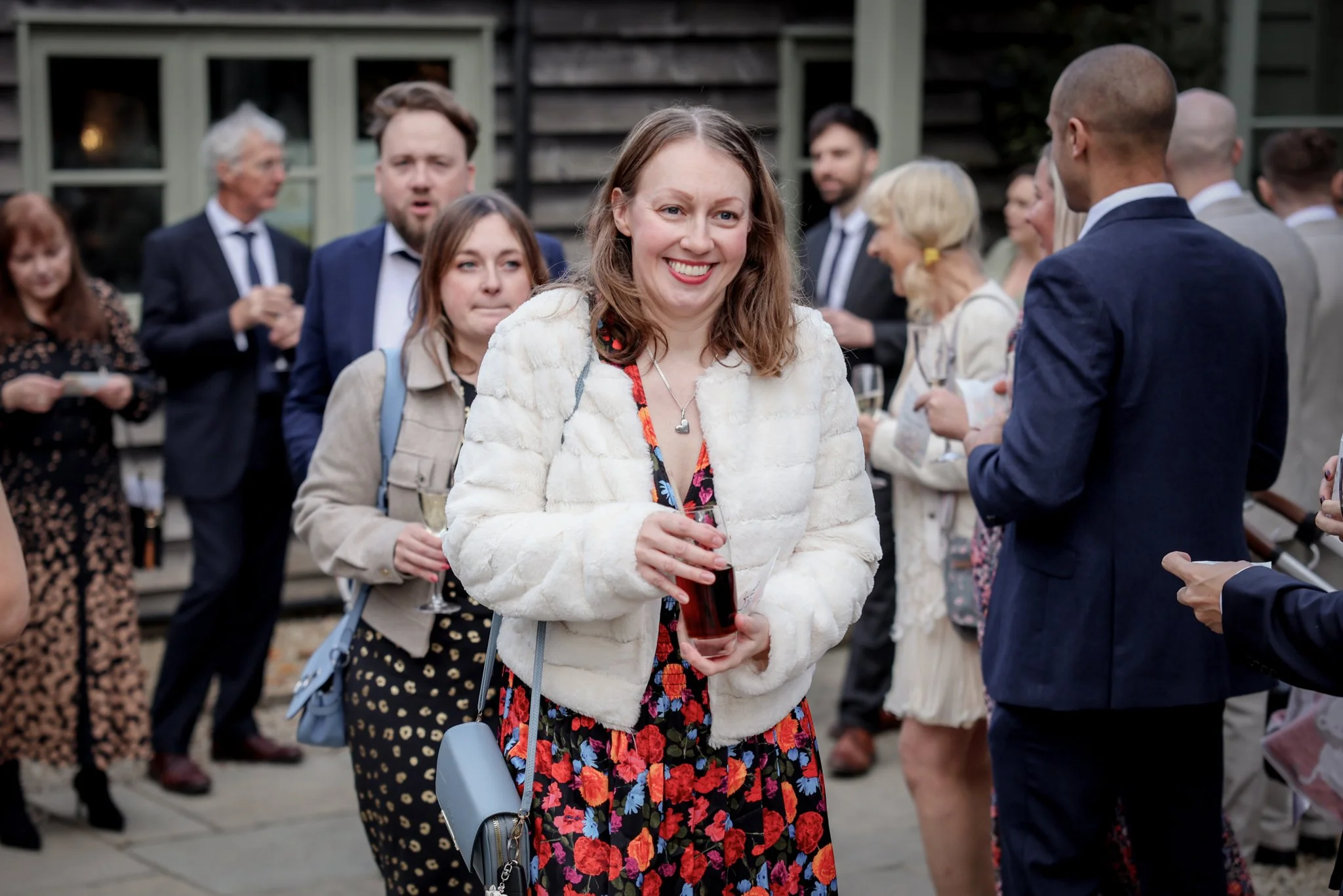 A group of people socializing outdoors at an event, with a woman in a white jacket and colorful dress smiling and holding a glass of wine in the foreground.