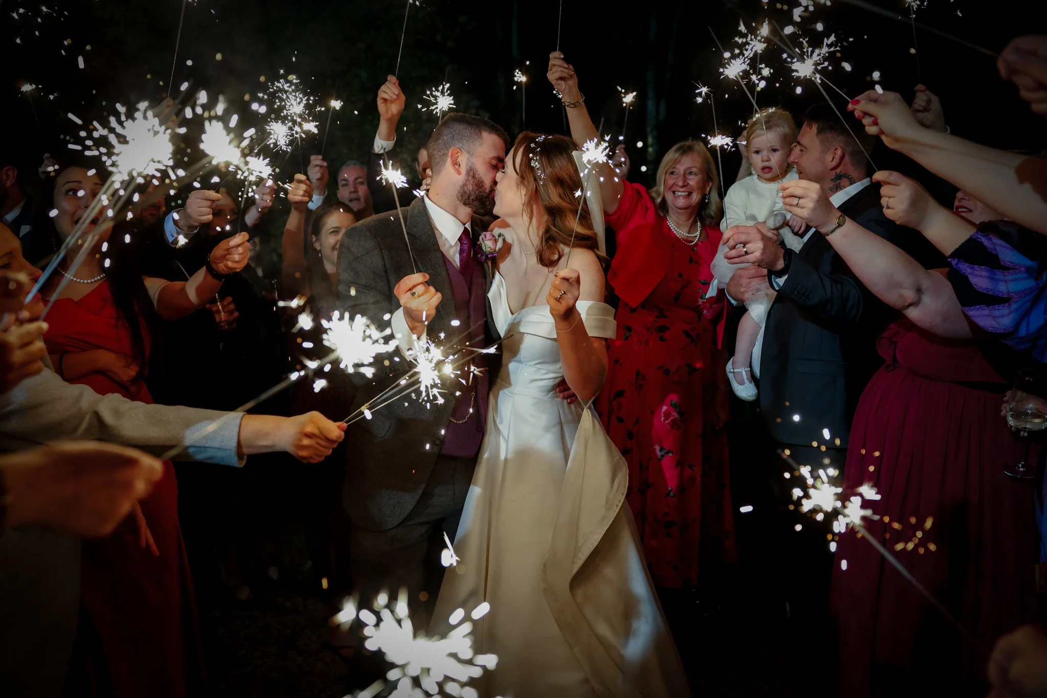 A bride and groom sharing a kiss during their wedding celebration surrounded by guests holding sparklers at night.