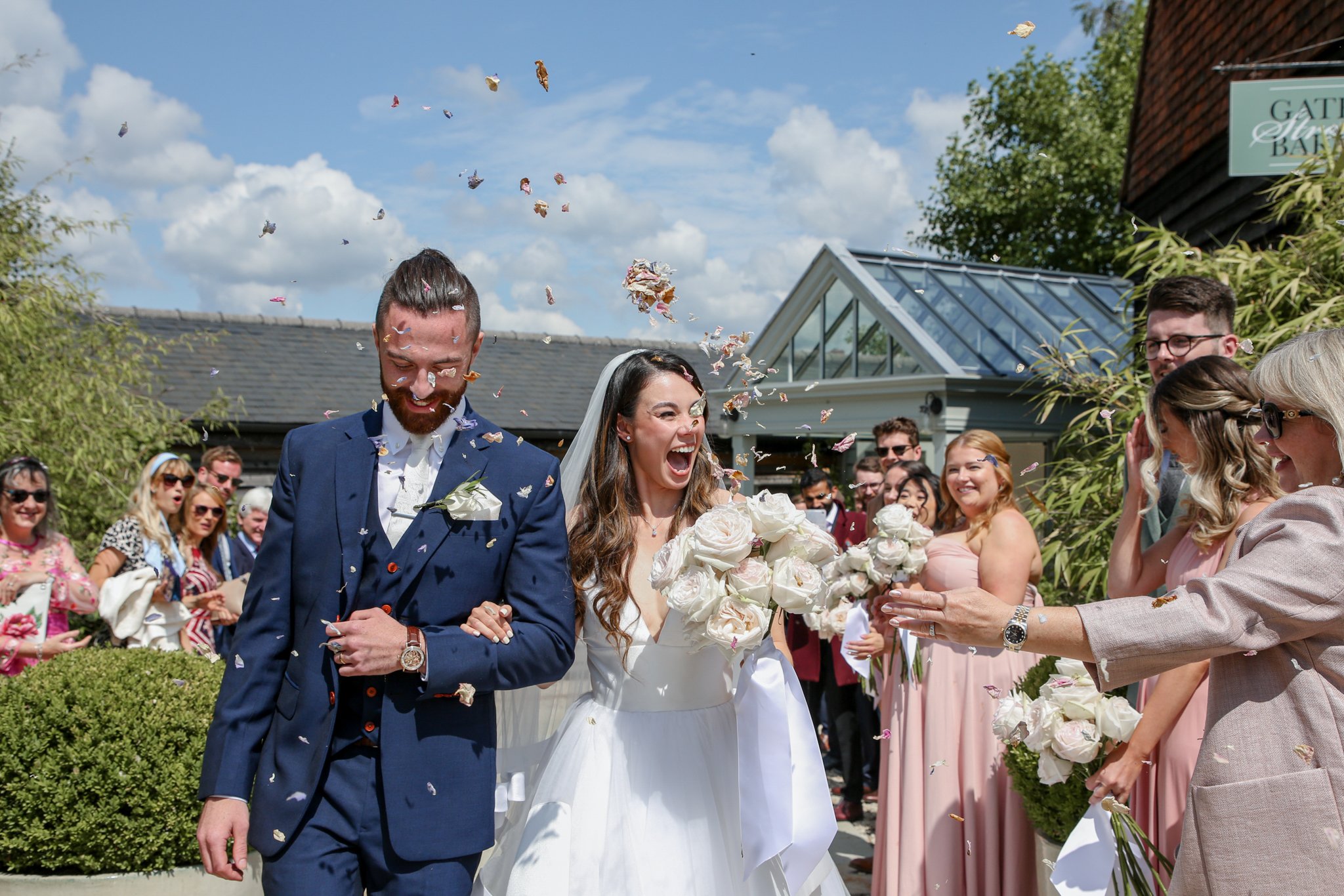 A bride and groom celebrating their wedding outdoors as guests throw flower petals in the air.